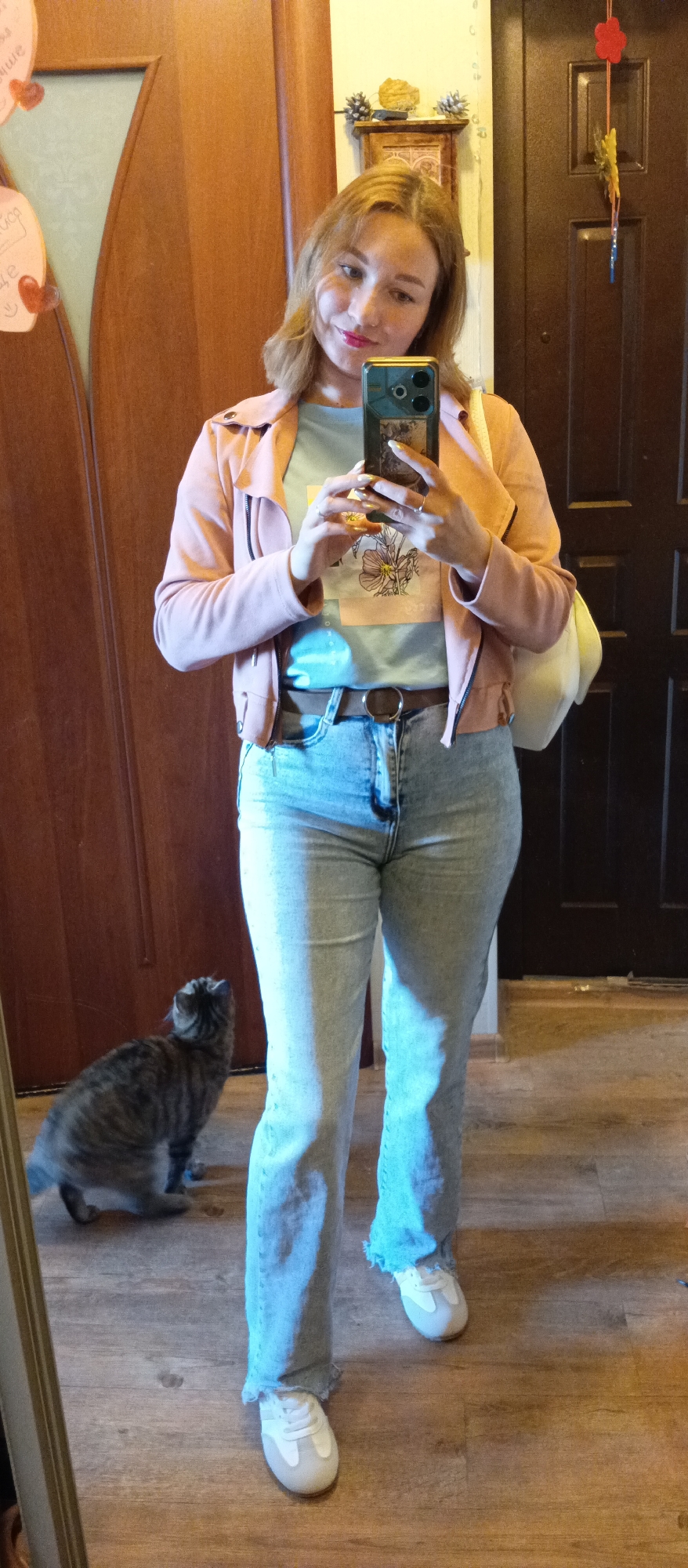 A young woman is taking a mirror selfie indoors. She is wearing a pink jacket, a blue t-shirt, blue jeans, and white sneakers. She is holding a smartphone in her hands, which is reflected in the mirror. Behind her to the left is a grey tabby cat looking up at her. A wooden door is to her right. The floor is brown wood. There is a shelf above the door with decorative items.