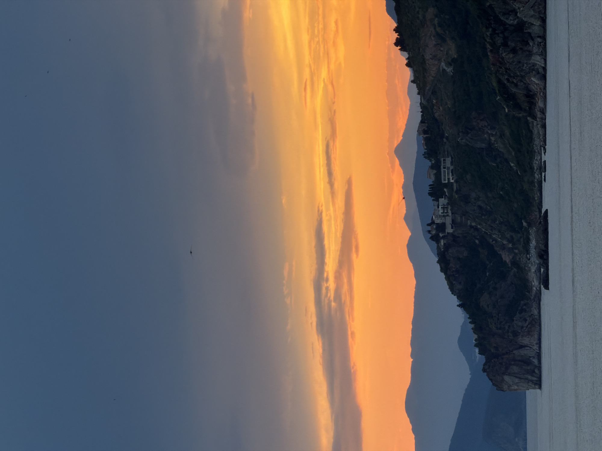 This is a landscape photograph taken at sunset in Petrovac na Moru, Montenegro. The image is oriented vertically, with the sky dominating the left side and the land and sea on the right. The sky displays a vibrant gradient of colors, transitioning from a deep blue at the top to fiery oranges and yellows near the horizon, indicative of a sunset. Wispy clouds are scattered across the sky, catching the last rays of sunlight.
In the mid-ground, a rugged, dark green mountainside slopes down towards the sea. Several white buildings, likely houses or villas, are visible nestled into the hillside, suggesting a coastal settlement. The texture of the mountain is rough and rocky, with patches of vegetation.
The foreground features the calm, greyish surface of the sea. The water reflects the muted light of the sky, creating a serene atmosphere. In the distance, the silhouettes of more mountains are visible, fading into a hazy blue, adding depth to the scene. A few small, dark specks in the sky suggest birds in flight. The overall mood is peaceful and tranquil, capturing the beauty of a coastal sunset. The time of day is clearly sunset, and the weather appears calm with clear skies except for the clouds.