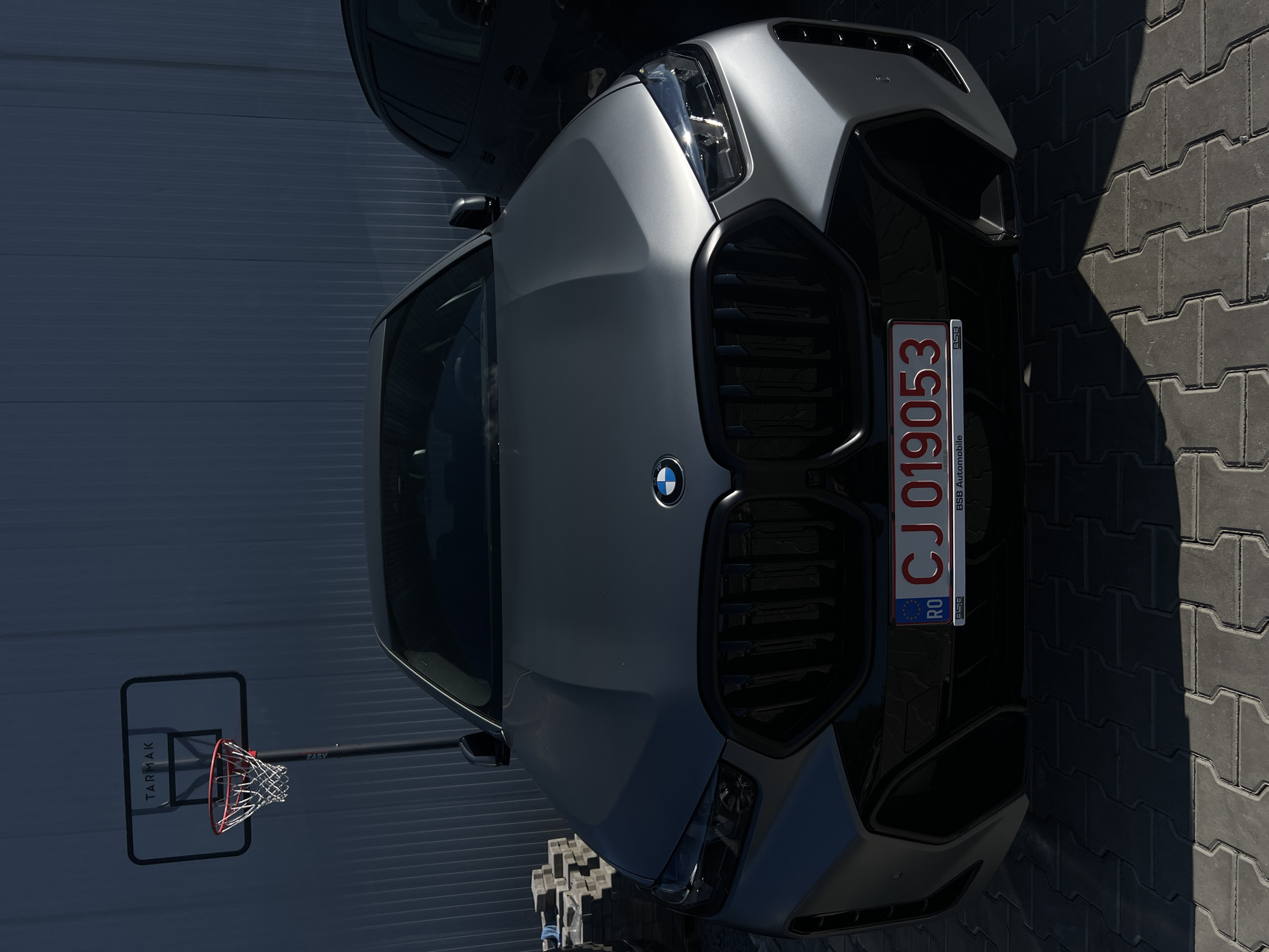 A gray BMW SUV is parked on a cobblestone surface in front of a dark blue corrugated metal wall. A basketball hoop with the "TARMAK" logo is visible to the left of the vehicle. The car has a Romanian license plate that reads "CJ 019053". The license plate frame also shows "BSB Automobile". The scene appears to be in an outdoor urban setting, possibly a parking lot or driveway.