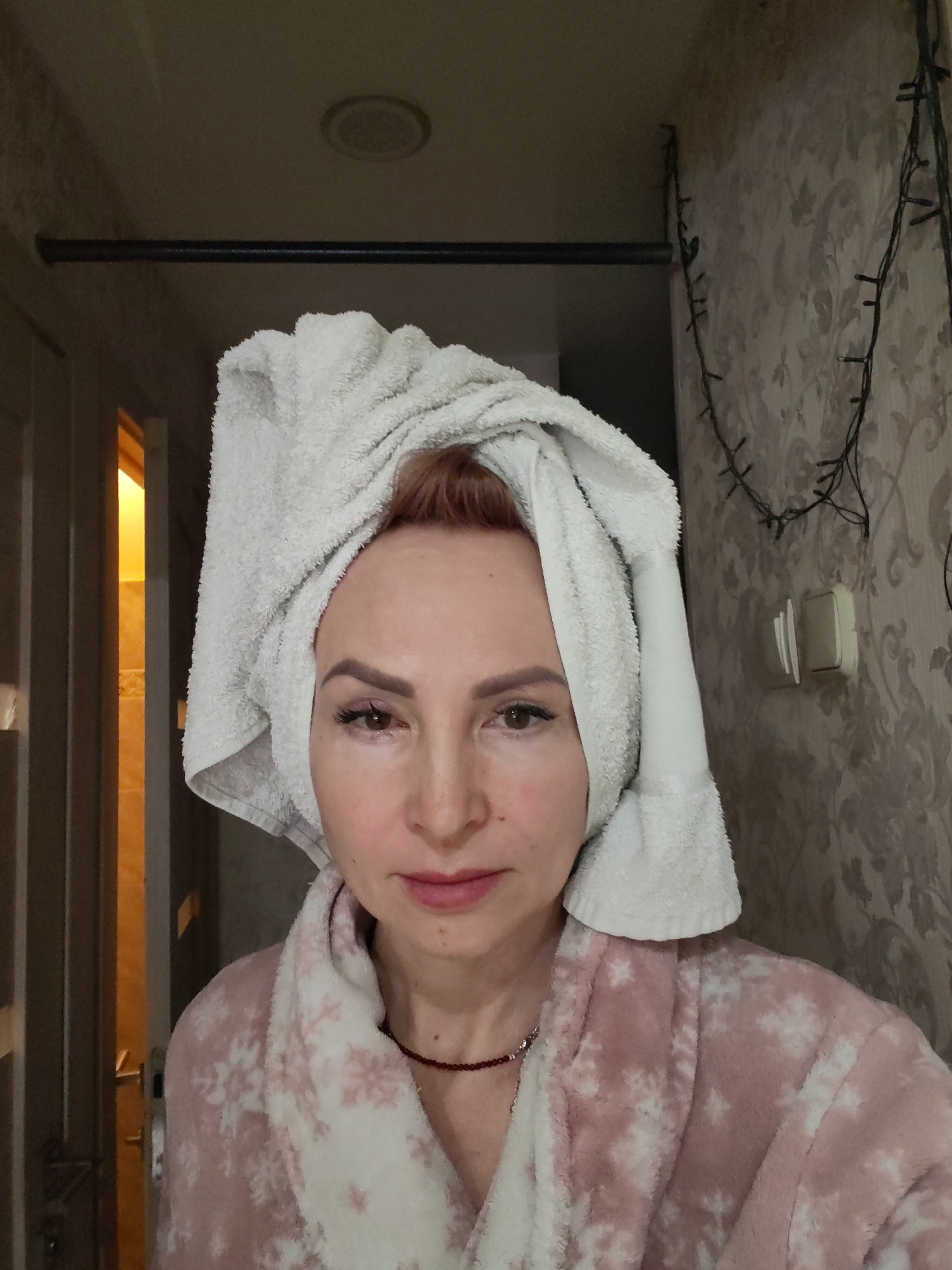 A forensic description of the image, located in the city of Chaikovsky, Russia, reveals a static indoor scene. The primary subject is a Caucasian woman, mid-shot, facing directly forward with a neutral expression. Her reddish-brown hair is wrapped in a white terrycloth towel styled as a turban. She is wearing a soft pink and white patterned bathrobe, likely made of fleece or terrycloth, featuring a snowflake-like design. A slender red beaded necklace with a small metallic element is visible around her neck.
The background consists of a room interior. To the woman's left, a dark-paneled interior door is slightly ajar, revealing a warmly lit room beyond, with light-colored tiled walls partially visible. A silver-toned door handle is discernible on the door. Above this, a dark horizontal bar or rod is mounted. To the woman's right, the wall is covered in light-toned patterned wallpaper with a subtle floral or damask design. Two light switches or electrical outlets are affixed to this wall. Two strands of small, dark string lights, resembling Christmas lights, hang vertically on the wallpapered wall. The ceiling is plain and features a recessed circular light fixture or vent.