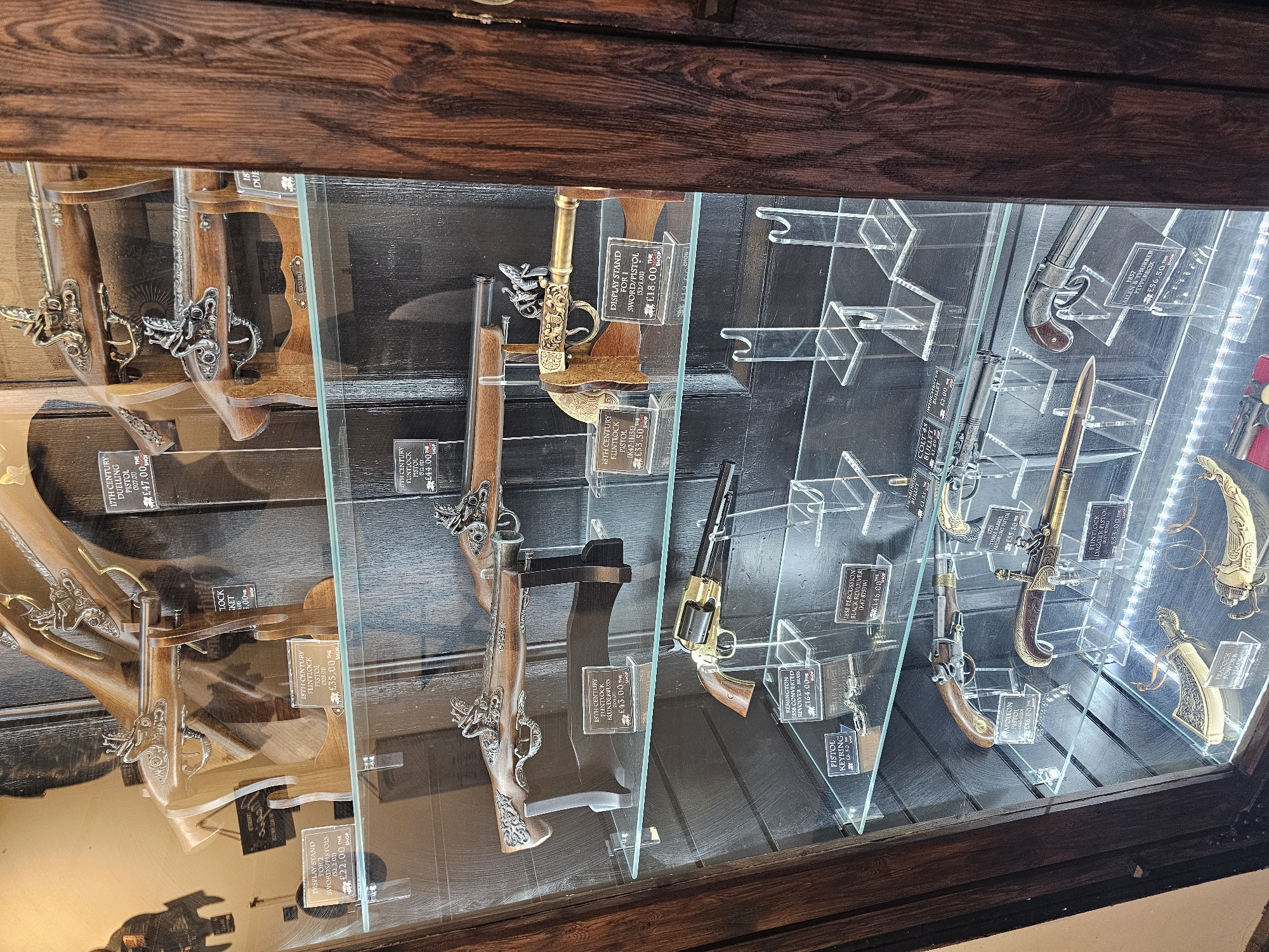 A display case with antique firearms is located in a store, likely a historical or souvenir shop, in Llandudno Junction, United Kingdom. The firearms include flintlock pistols, blunderbusses, and revolvers. Each firearm is displayed with a price tag and description, such as "18th Century Flintlock Pistol" or "1858 Percussion Black Revolver." The display case is made of wood and glass, and the firearms are arranged on acrylic stands. Some stands also display display stand, pistol keyrings, a Colt's bullet, and other items. The pricing shown indicates that these are probably reproductions.