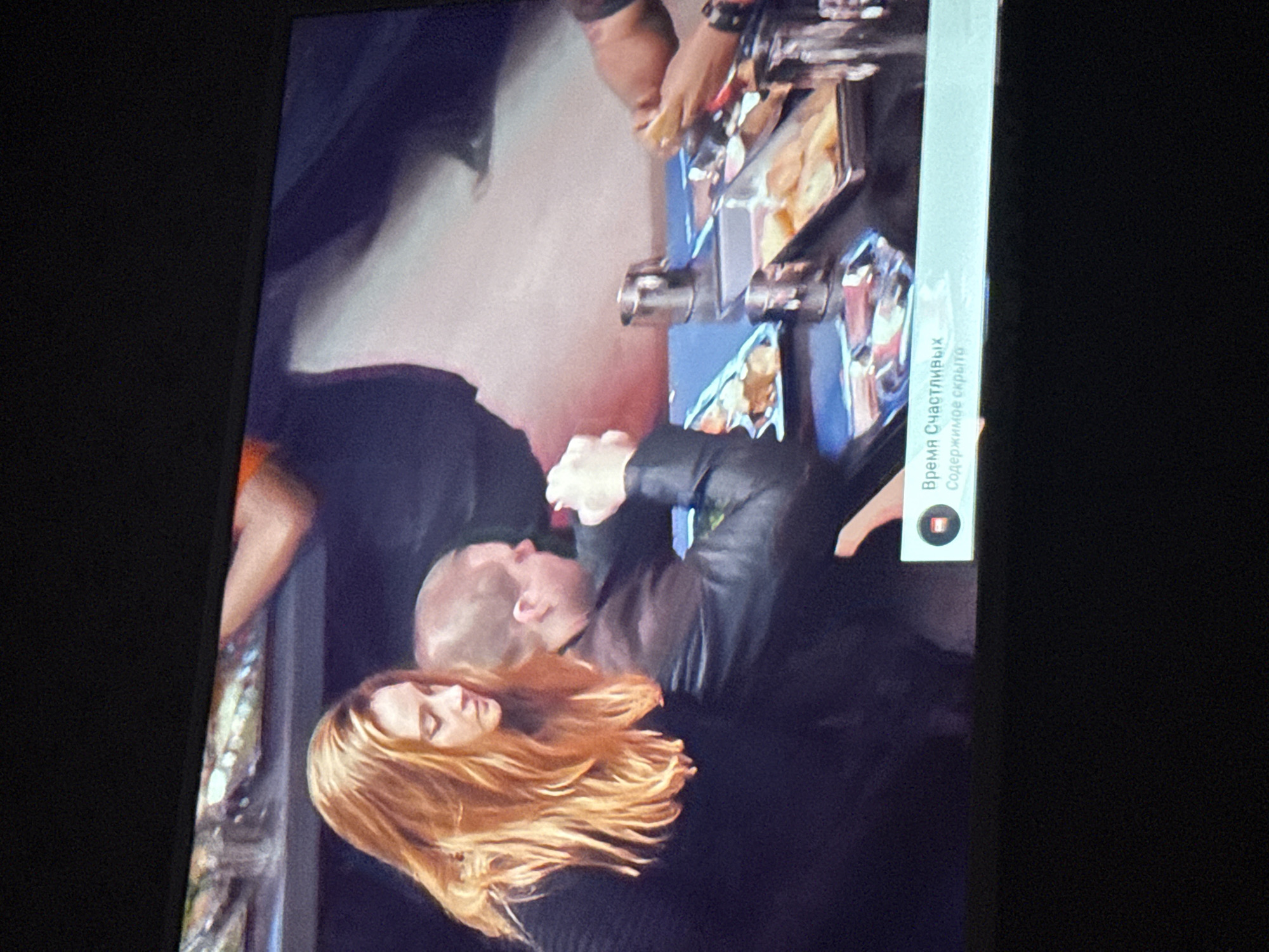 A medium shot of a television screen. The screen shows a scene of two individuals standing. The woman has long, reddish-blonde hair and is wearing a dark top. A bald-headed man in a dark jacket stands next to her, with his hands in a folded position. They are seemingly at an event where food and drinks are being served, with a person in the upper portion of the frame appearing to be engaged with serving or preparing refreshments. A notification, written in Russian, is visible in the top-right corner of the screen. The location information (Mytishchi, Russia) cannot be confirmed from the image content alone.