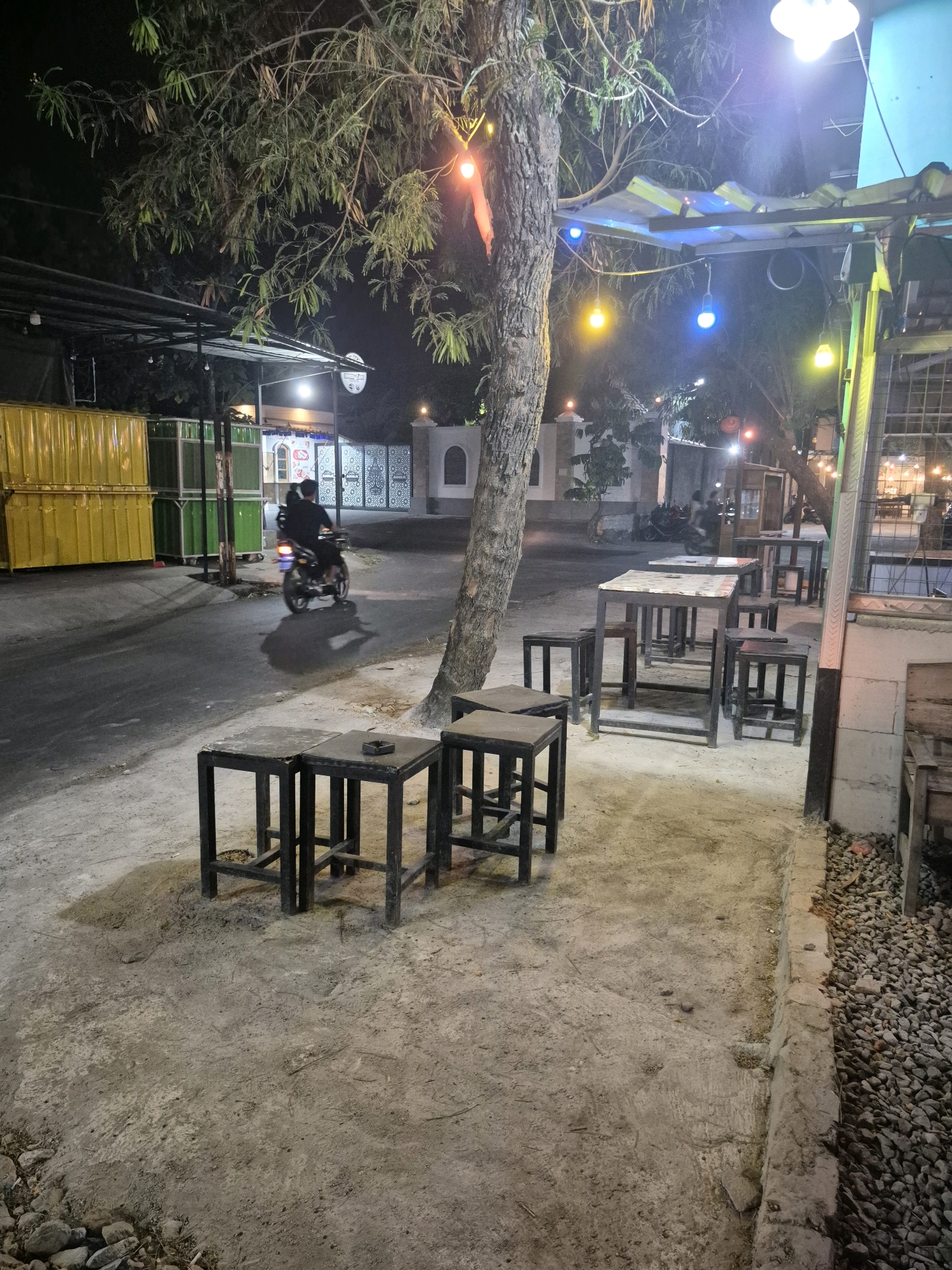 The image depicts an outdoor seating area at night in Pabelan, Indonesia, likely a cafe or restaurant. A motorcycle with a rider is seen on the road in the background. Several small tables and stools are arranged on a dirt and concrete ground. String lights with colored bulbs are visible, illuminating the scene along with a bright overhead light. The area appears to be adjacent to a street with some commercial buildings or containers in the background. The atmosphere is casual and slightly rustic.