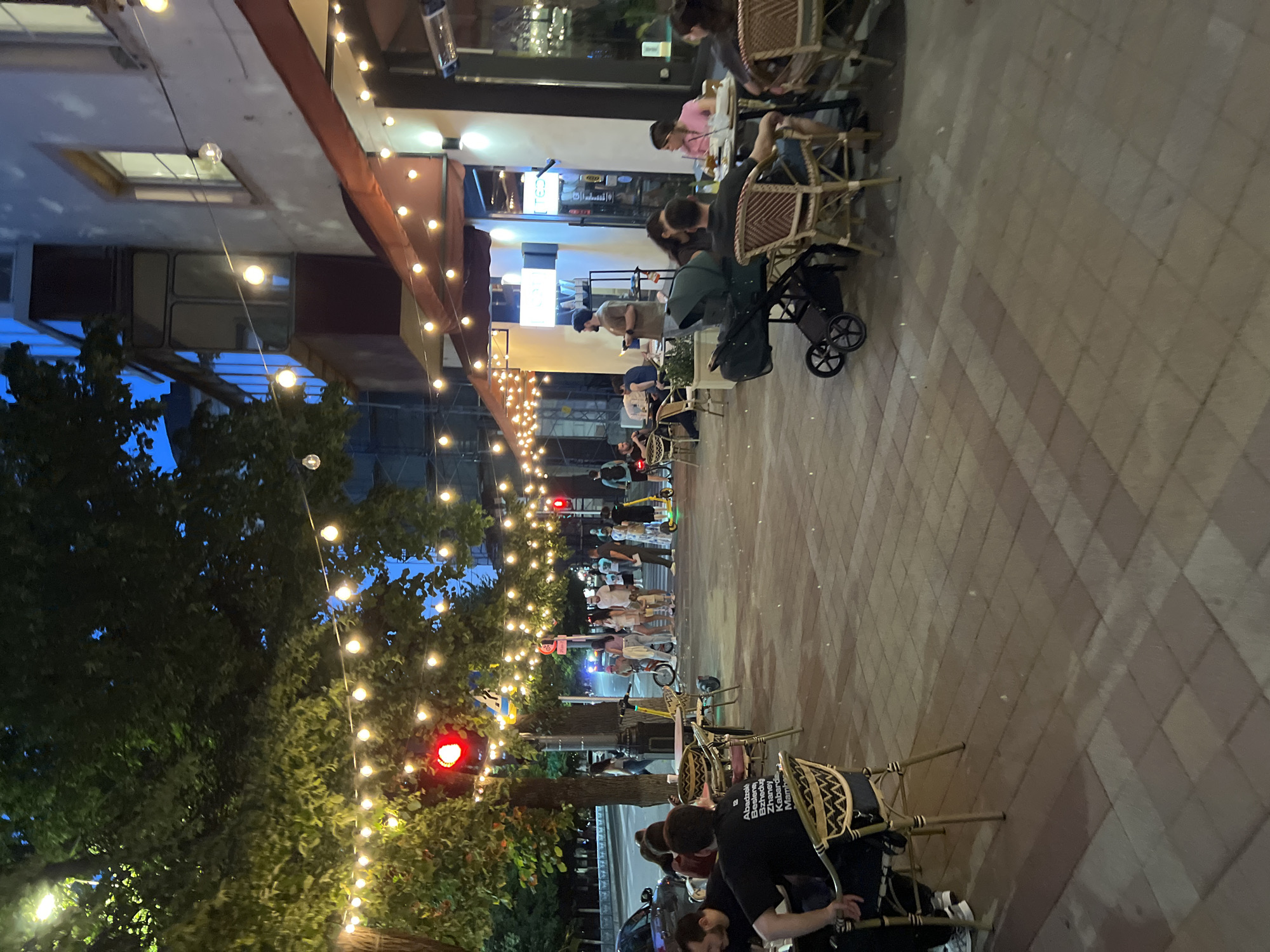 The image depicts an outdoor dining area in Krasnodar, Russia, at dusk. String lights illuminate the scene, casting a warm glow on the paved walkway and tables. Several people are seated at outdoor tables, some appearing to be dining and conversing. A baby stroller is visible near one of the tables. In the background, a street with traffic lights and a few pedestrians crossing the street can be observed. The building adjacent to the dining area has windows and awnings, suggesting it is a cafe or restaurant. The presence of trees and the overall ambiance suggest a lively urban evening.