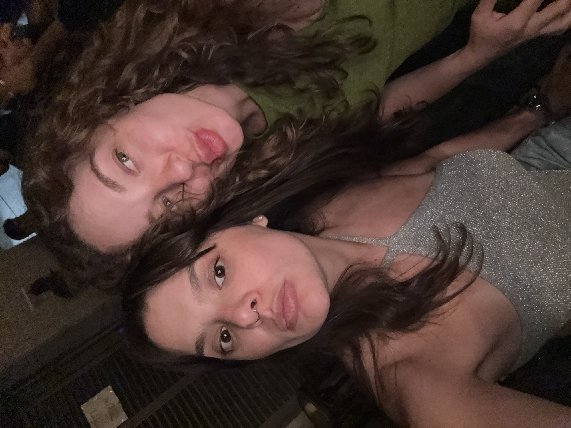 Two women are taking a selfie in a dimly lit indoor environment, possibly a bar or restaurant in Tel-Aviv, Israel. The woman on the left has dark brown hair, brown eyes, and a nose ring. She is wearing a sparkly silver top. The woman on the right has curly brown hair and is wearing a green top. She is puckering her lips for the photo. The background is dark with some blurred figures and what appears to be a sign on the left. The overall lighting is low, with flash illuminating the faces of the two women.