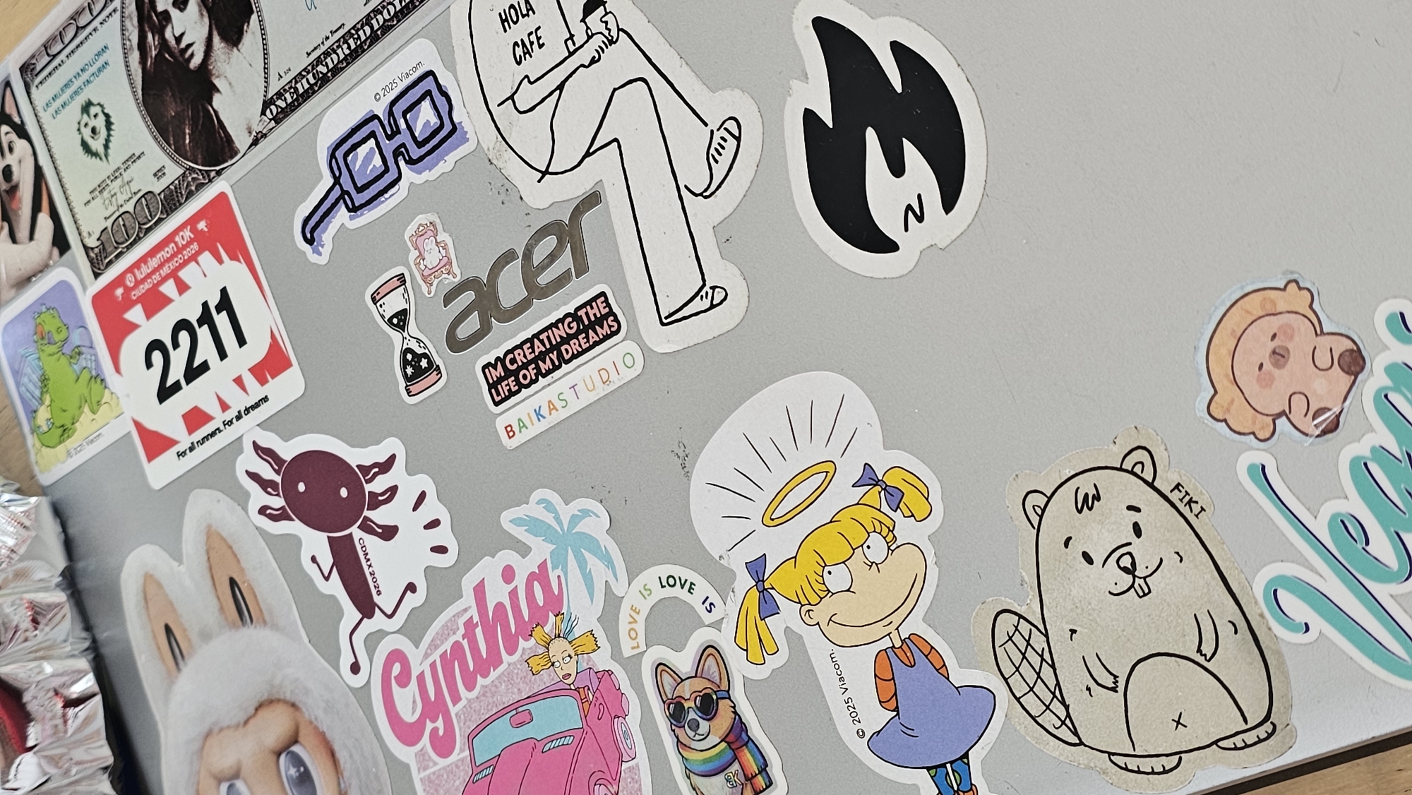 The image shows a laptop lid covered in stickers. Stickers include references to "Las Mujeres Ya No Lloran" and "Las Mujeres Facturan", a Lululemon 10K in Ciudad de Mexico (2026) with bib number 2211, an Acer logo with the text "I'm creating the life of my dreams", a "Hola Cafe" sticker, characters from Rugrats (Angelica and Cynthia), a beaver sticker with "FIKI" printed on it, and an axolotl sticker labeled "CDMX 2026".