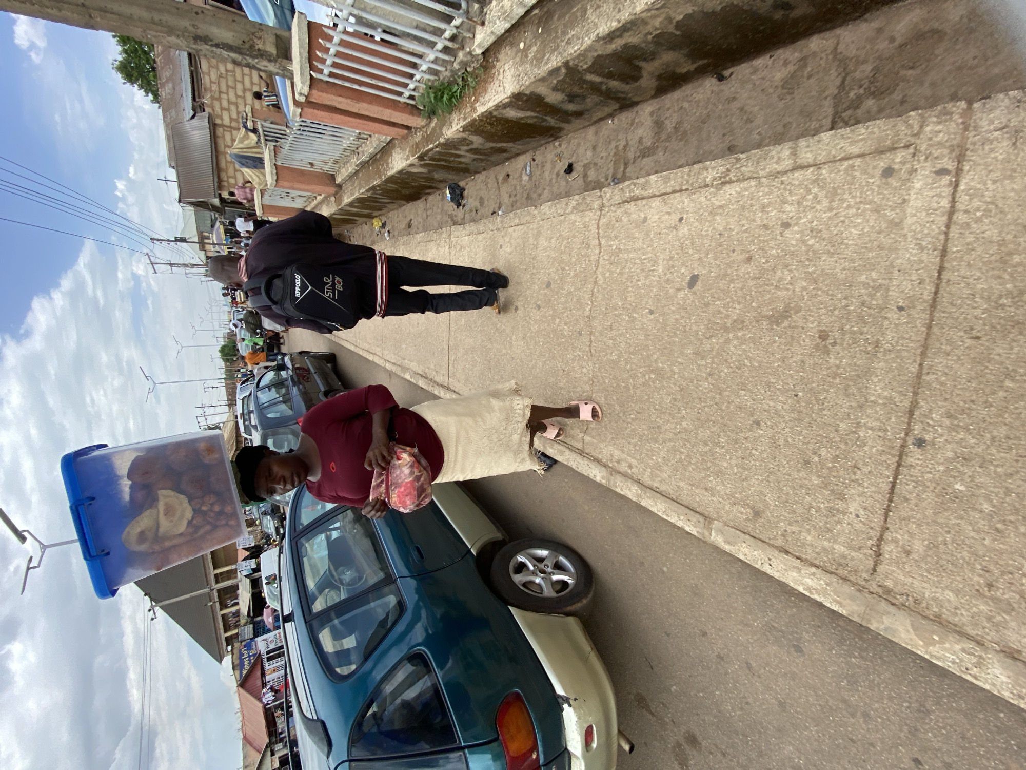 A street scene in Bukuru, Nigeria, features a woman carrying a large transparent container of food items. She wears a maroon top and tan skirt, and stands near a parked teal car. A pedestrian in a black jacket walks on the sidewalk. The street is lined with cars and shops. A white metal fence runs along the edge of the sidewalk. The sky is partly cloudy.