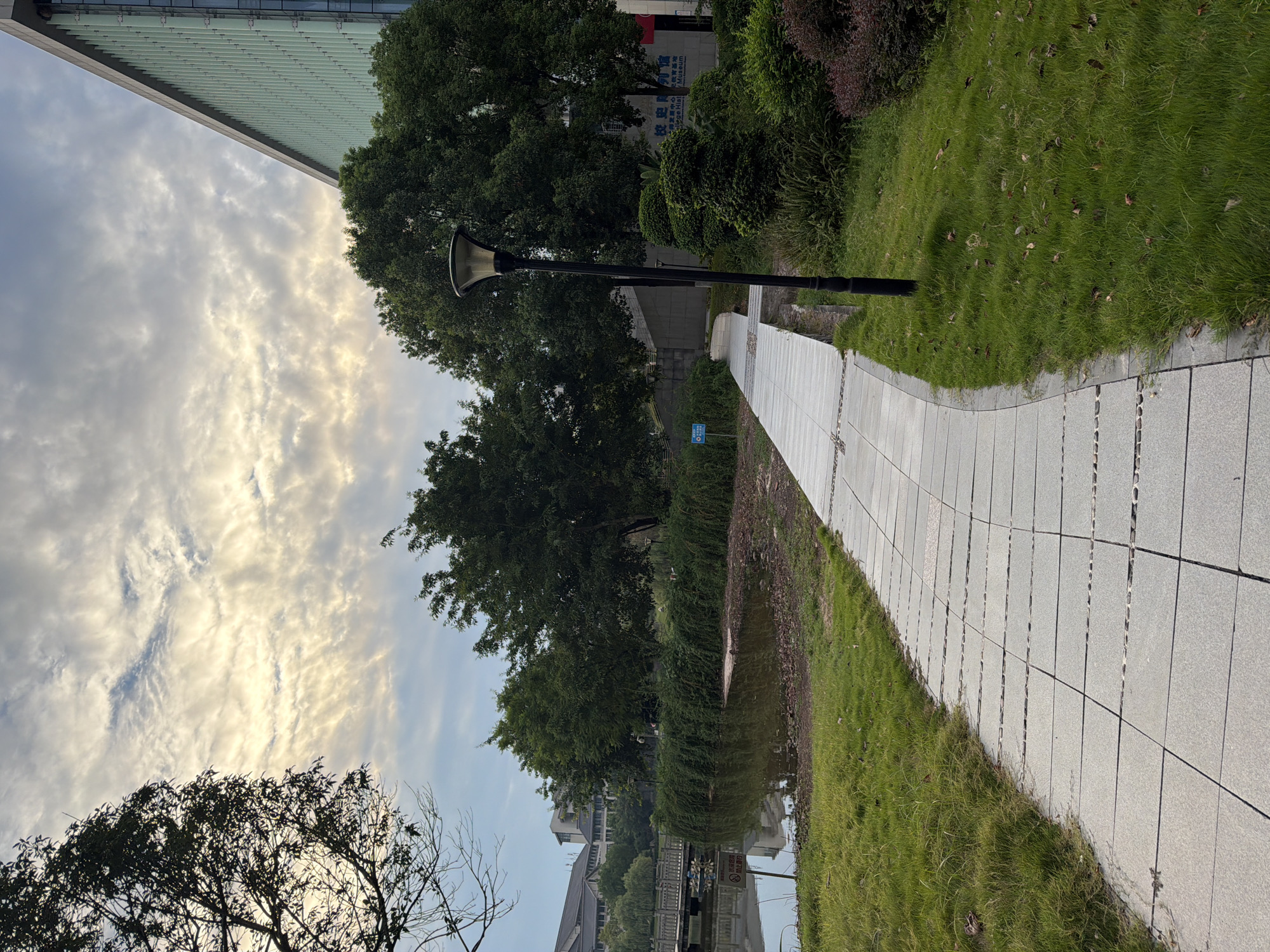 The image depicts an outdoor scene in a park-like setting, possibly on the grounds of a university or research facility in Jiangjin, China, judging by the architectural style and signage visible on the building in the background. A paved walkway, made of square grey tiles with pebble inlays, curves through the scene. To the left of the walkway is a body of water, likely a pond or small lake, with a wooden bridge in the distance. The water's edge is lined with dense green bushes and trees. To the right of the walkway, a neatly trimmed lawn is interspersed with ornamental shrubs and a black lamppost. A large, modern building with a green-tinted glass facade dominates the background on the right side. The sky is partly cloudy, with streaks of light suggesting either sunrise or sunset. No people are visible in the frame, and there are no evident actions or interactions taking place. The overall atmosphere is calm and serene.