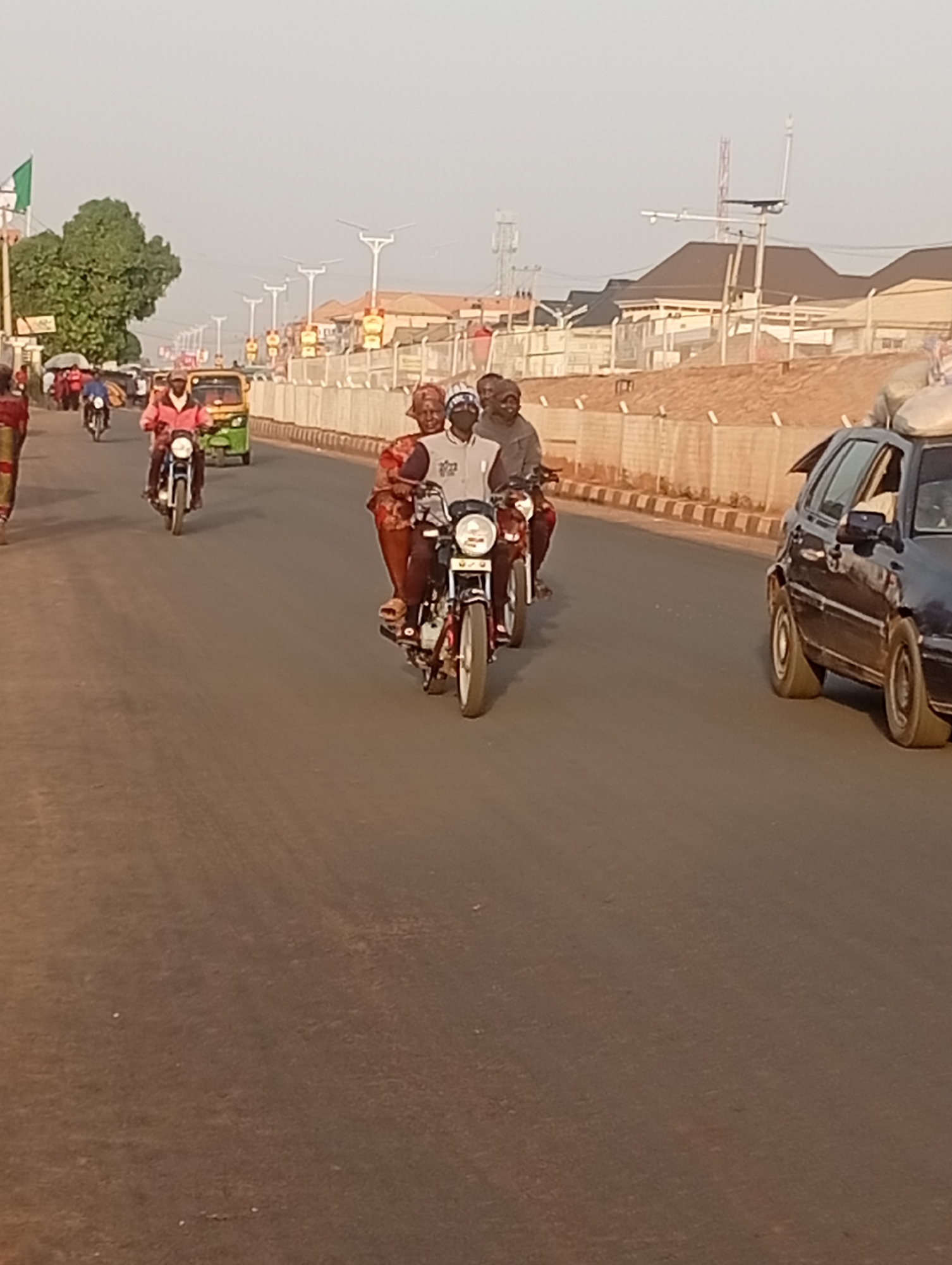 The image shows a street scene in Sabon Tasha, Nigeria. The street is paved and appears to be a main thoroughfare. In the foreground, a motorcycle carries three people: a driver, a woman behind him, and a man behind her. A second motorcycle follows closely behind with a single rider. To the left, a person walks along the side of the road, and a three-wheeled vehicle (a keke) and another motorcycle are farther ahead. To the right of the motorcycles, a dark-colored car with items strapped to the roof is moving in the same direction. The background reveals buildings, possibly residential or commercial, set behind a low barrier. Streetlights and utility poles are visible. The sky is overcast, suggesting daytime.