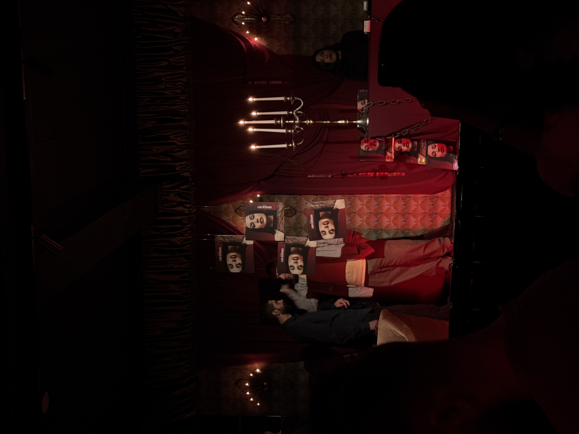 The image depicts a darkly lit indoor performance or event space, likely a stage area, in Yerevan, Armenia, consistent with the aesthetic of venues such as Aeon. The environment features a backdrop of red velvet drapes and dark, textured wallpaper with a repeating, possibly scale-like pattern. Ornate, multi-branched wall sconces with small light bulbs illuminate the background on both sides.
In the center-left, a bearded man in a dark shirt stands, facing towards the right. To his immediate right, another individual wears a light-colored plaid shirt and a red vest or scarf, standing near a display of hanging square posters. These posters prominently feature a woman's face and bear the text "LOVE REVERSED." On the right side of the stage, a tall, silver-colored candelabra with five lit candles stands on a dark red table or platform. Behind this table, a woman with dark hair, wearing a black top, stands observing the scene. Dark metal chains are draped across the red table. Below the table, additional vertically stacked images of a woman's face are visible. The foreground shows the silhouettes of audience members, indicating an ongoing event.