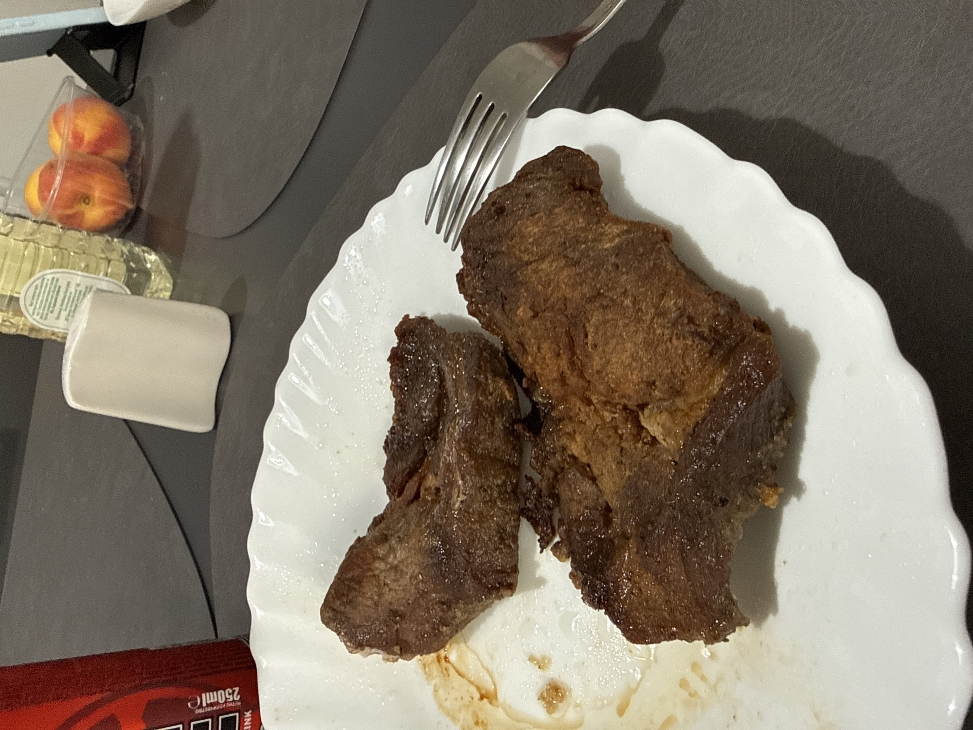 The image depicts a white, scalloped-edged plate containing two pieces of cooked meat, and a fork resting on top of the larger piece. The meat appears to be browned and possibly seasoned. Traces of residue, possibly sauce, are visible on the plate beneath the meat.
Additional objects are partially visible around the plate including a container of peaches, a white container and a red and black box. The location is not ascertainable from the image. Stara Zagora, Bulgaria, cannot be verified or dismissed.