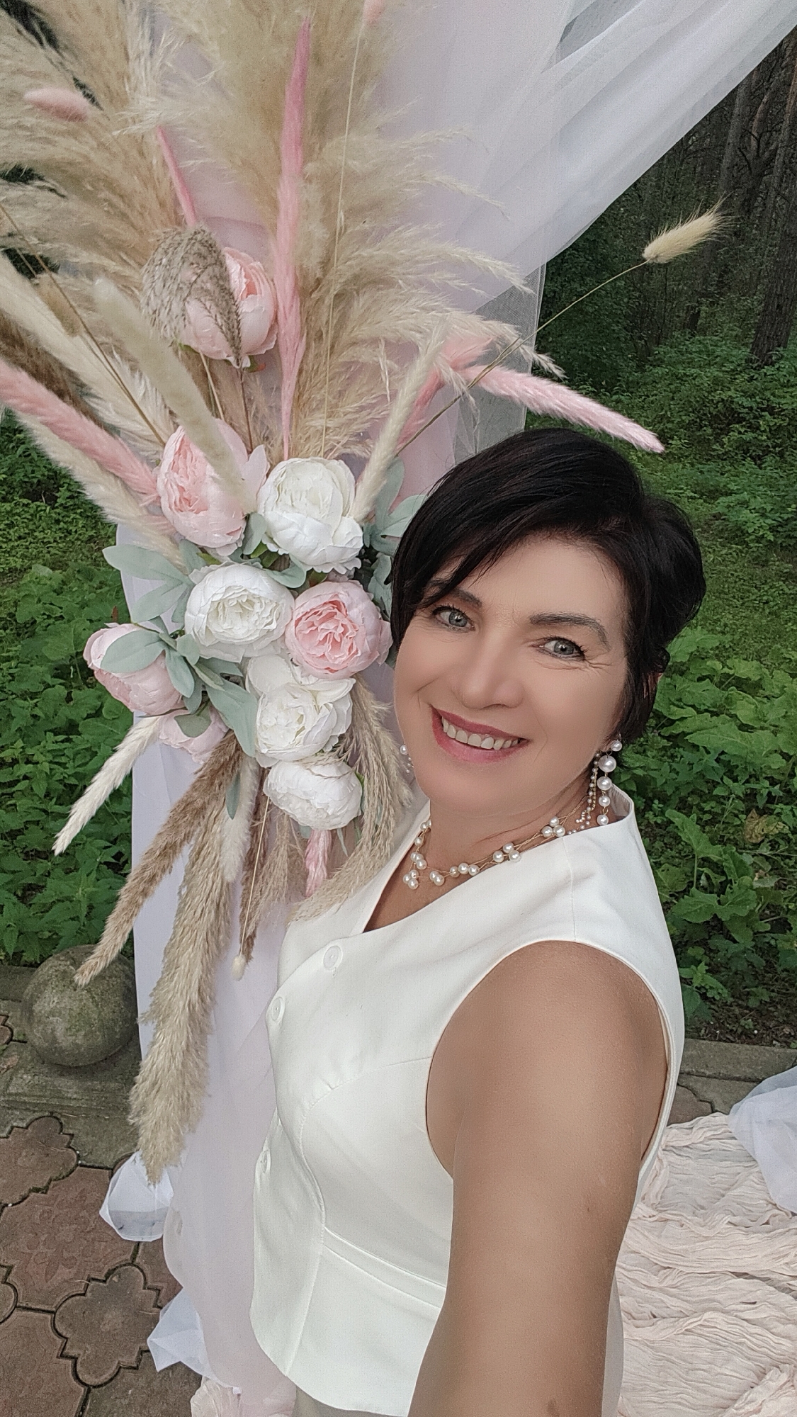 A woman with short, dark hair and blue eyes is standing outdoors, smiling at the camera. She is wearing a white sleeveless vest and a pearl necklace. Behind her is a floral arrangement of light pink and white peonies, along with dried pampas grass and other decorative elements, set against a backdrop of greenery and trees. A white sheer fabric drapes from an unseen structure above. The ground beneath her is paved with decorative stone tiles. The location is identified as the city of Beryozovka, Russia.