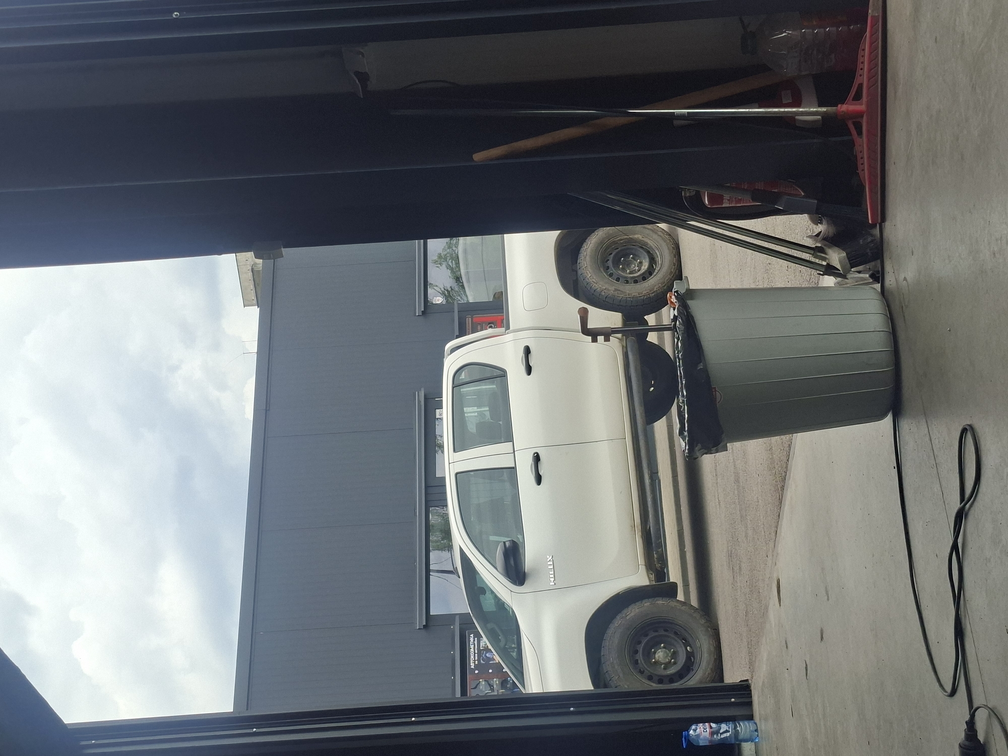 Observed from within a covered structure, this scene depicts a white Toyota Hilux parked next to a gray trash can on a concrete surface. A gray building is visible behind the truck. The area appears to be a garage or storage space, with various objects like a broom, bottles, and power cords scattered around. The sky is visible through the opening. The city and location are unknown.
