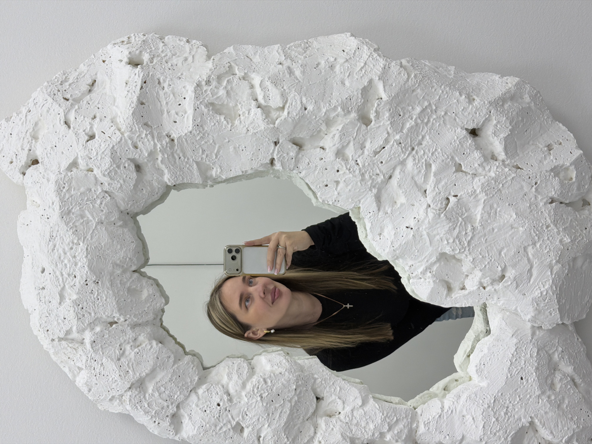 A woman captures her reflection in a selfie taken in a mirror with a distinctive, heavily textured white frame. The frame appears to be made of a rough, porous material, resembling raw concrete, coral, or abstract plasterwork, creating an irregular and organic border around the reflective surface.
In the mirror, the woman is seen from the chest up, positioned at an angle as she holds a light-colored smartphone with her right hand to take the photograph. She has long, straight, brown hair with lighter highlights, blue eyes, and a calm, slightly upward-gazing expression. She wears a black long-sleeved top, a delicate gold necklace with a cross pendant, and a small pearl earring in her visible ear. The background reflected in the mirror is a plain white wall. The overall scene is brightly lit indoors. The provided context indicates the location is Moscow, Russia. There is no visible text in the image.