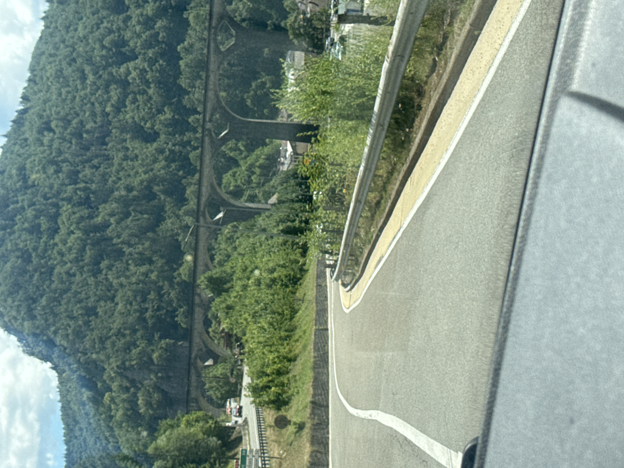 The image shows a view from a vehicle traveling on a winding road. The road has a white line marking the center and a yellow line marking the edge. To the right of the road is a metal guardrail and grassy embankment. In the background, a large, arched viaduct spans across a valley, with dense green forest covering the surrounding hills. A few buildings are visible beneath the viaduct. The sky is partly cloudy. The location is Hauts de Bienne, France.
