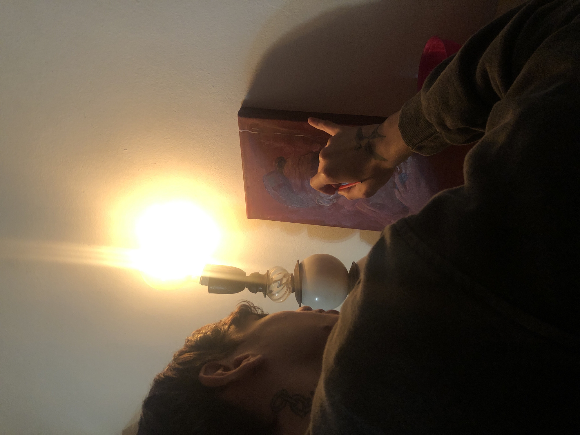 A person is painting a canvas propped against a wall indoors. The person is wearing a dark-colored sweatshirt and has tattoos on their hand and neck. A lamp is visible near the person's head, providing light for the painting. The canvas appears to have an abstract design. The location is indoors, but there is no indication of the city or country.