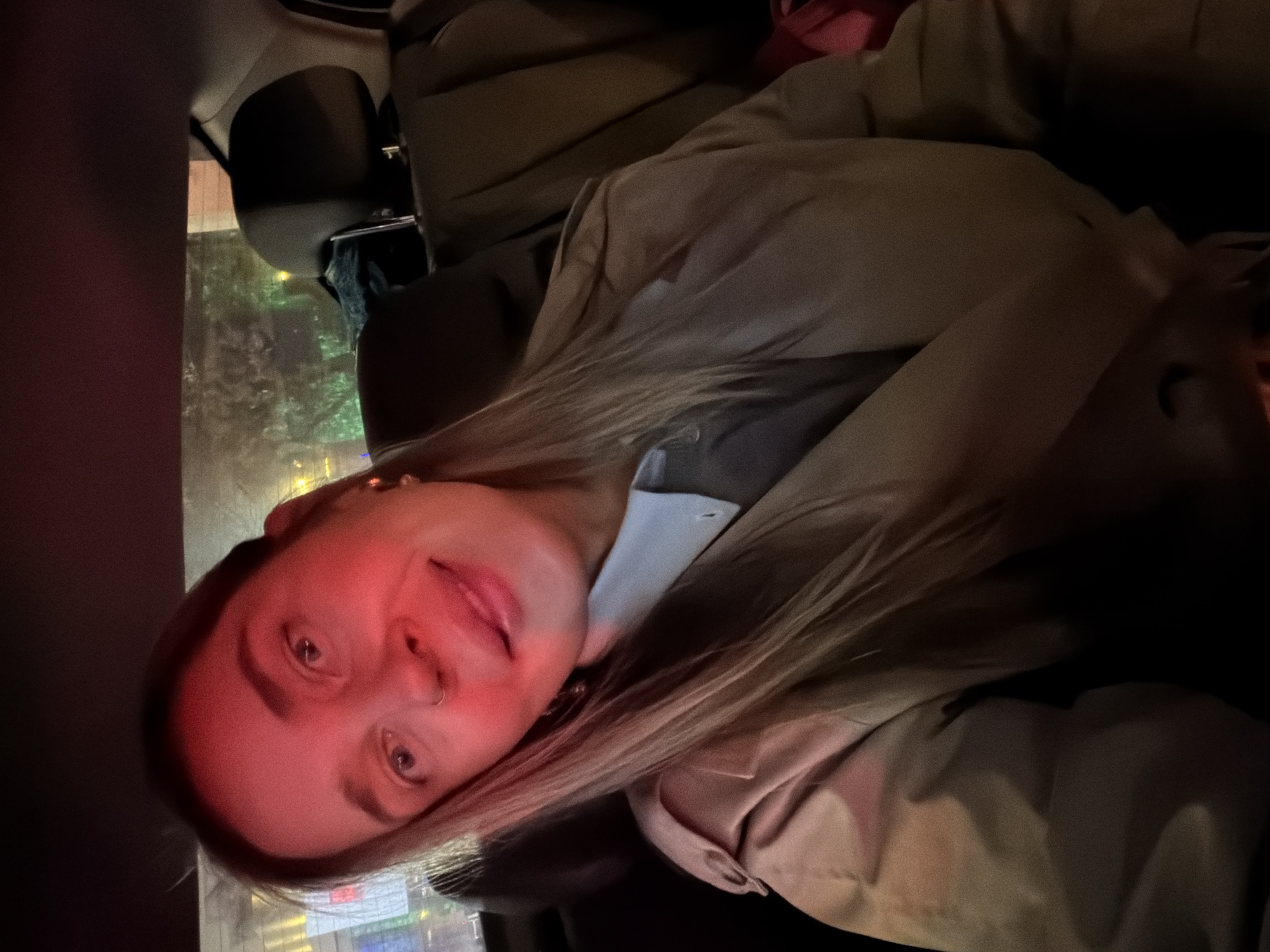 A woman with fair skin and long blonde hair is seated in a vehicle, likely a car, during nighttime in Moscow, Russia. She is wearing a light-colored jacket or coat and a pale shirt or blouse underneath. The ambient lighting casts a reddish hue on her face. Outside the car window, blurred city lights and illuminated buildings are visible, consistent with a Moscow cityscape. The interior of the car is dark, with portions of the seats and car interior visible around her. The overall atmosphere suggests a casual moment within the vehicle, possibly during travel through the city at night.