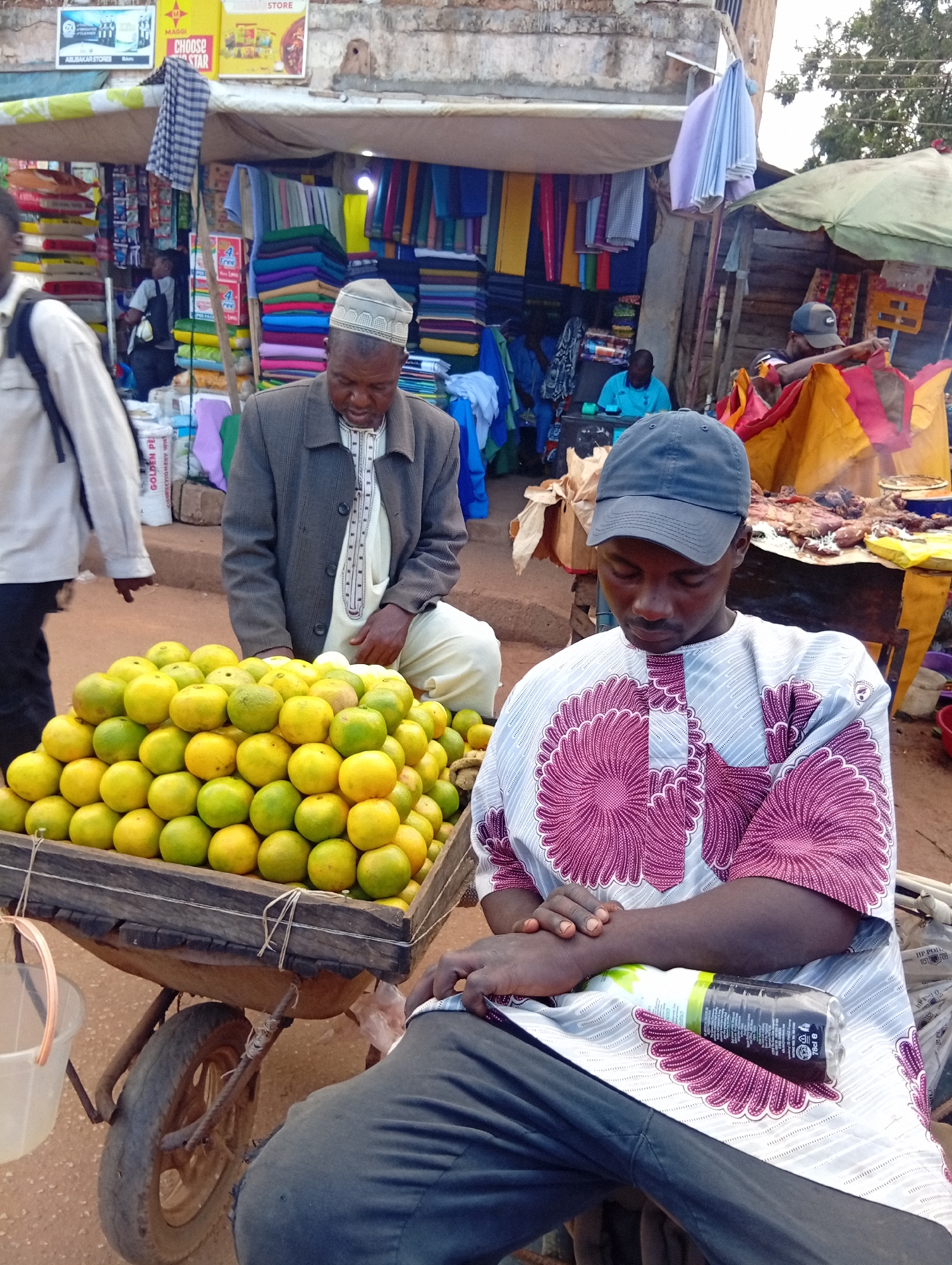 The image depicts a street market scene in Bukuru, Nigeria. In the foreground, a man wearing a grey jacket and traditional white attire with patterned trim is seated next to a large wooden cart filled with ripe oranges. His gaze is directed downwards, and his hands are clasped. Beside him, another man wearing a patterned traditional shirt and a dark baseball cap is also seated, looking downwards with his arms crossed. A plastic bottle lies on his lap.
Behind the seated men, a busy market stall is visible, featuring stacks of colorful textiles and fabrics. Several other individuals are present in the background, engaged in market activities. To the right, a food stall displays grilled meat. The overall environment suggests an outdoor marketplace with goods being sold from stalls and carts. The ground is composed of reddish-brown earth.