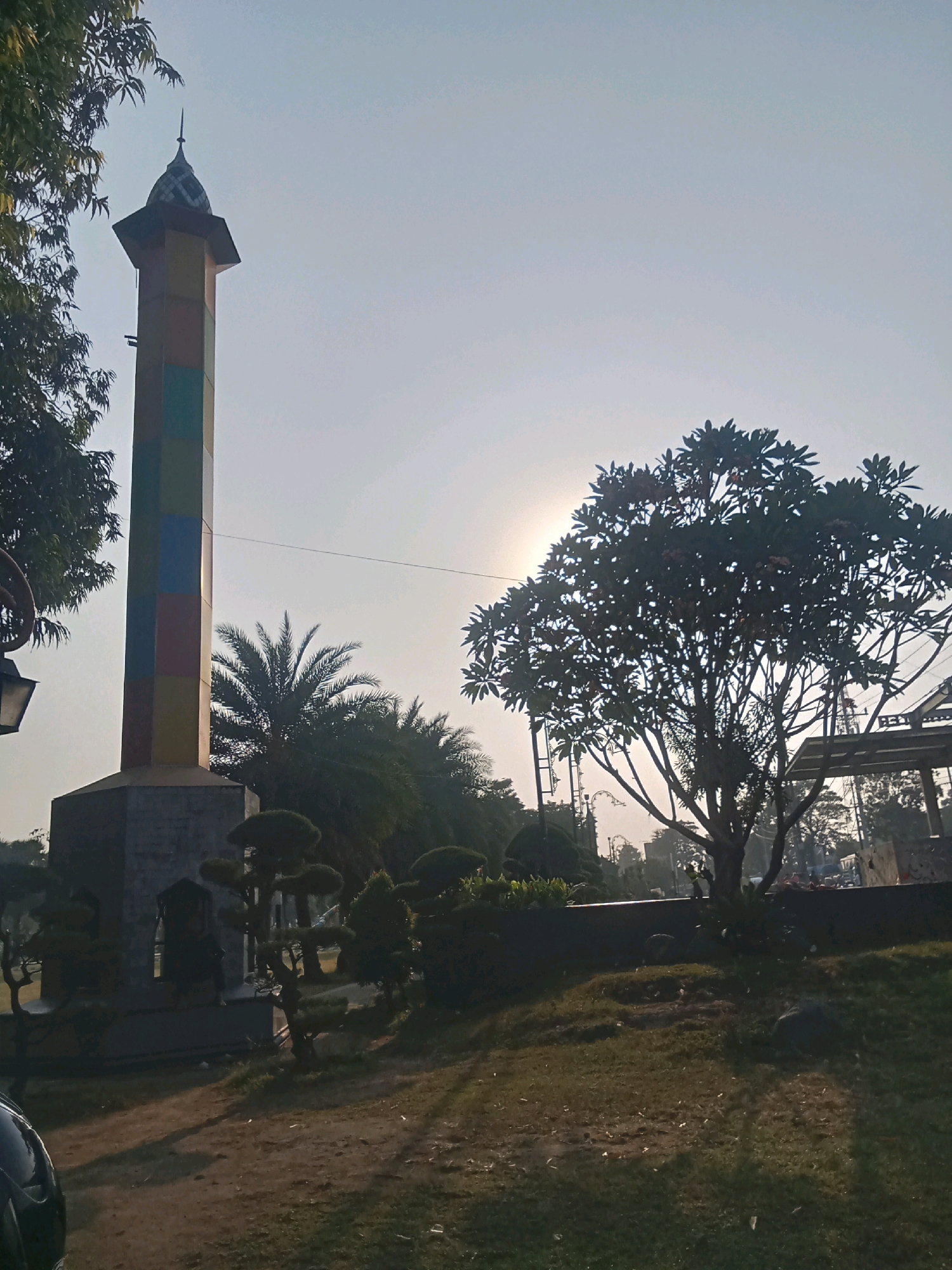 A tall, multi-colored obelisk stands prominently in a park setting under a bright, hazy sky, with the sun partially obscured by a large tree. The obelisk is segmented into horizontal bands of yellow, green, blue, and red, topped by a dark, pointed spire. At the base of the obelisk is a hexagonal stone structure.
To the right of the obelisk, a leafy tree with a wide canopy dominates the mid-ground, its silhouette stark against the sky. The sun's glare is visible through the branches of this tree. In front of the tree and around the obelisk, well-maintained bushes and small trees are visible, some appearing to be carefully pruned.
The park grounds are covered in grass and dirt. In the background, beyond the trees and structures, a hazy cityscape is suggested with various poles and wires, hinting at a suburban or urban environment. A glimpse of a dark-colored vehicle is visible in the lower-left corner of the frame. The overall impression is a daytime scene in a public park, possibly within the city of Kaliwungu, Indonesia, given the architectural style and vegetation.