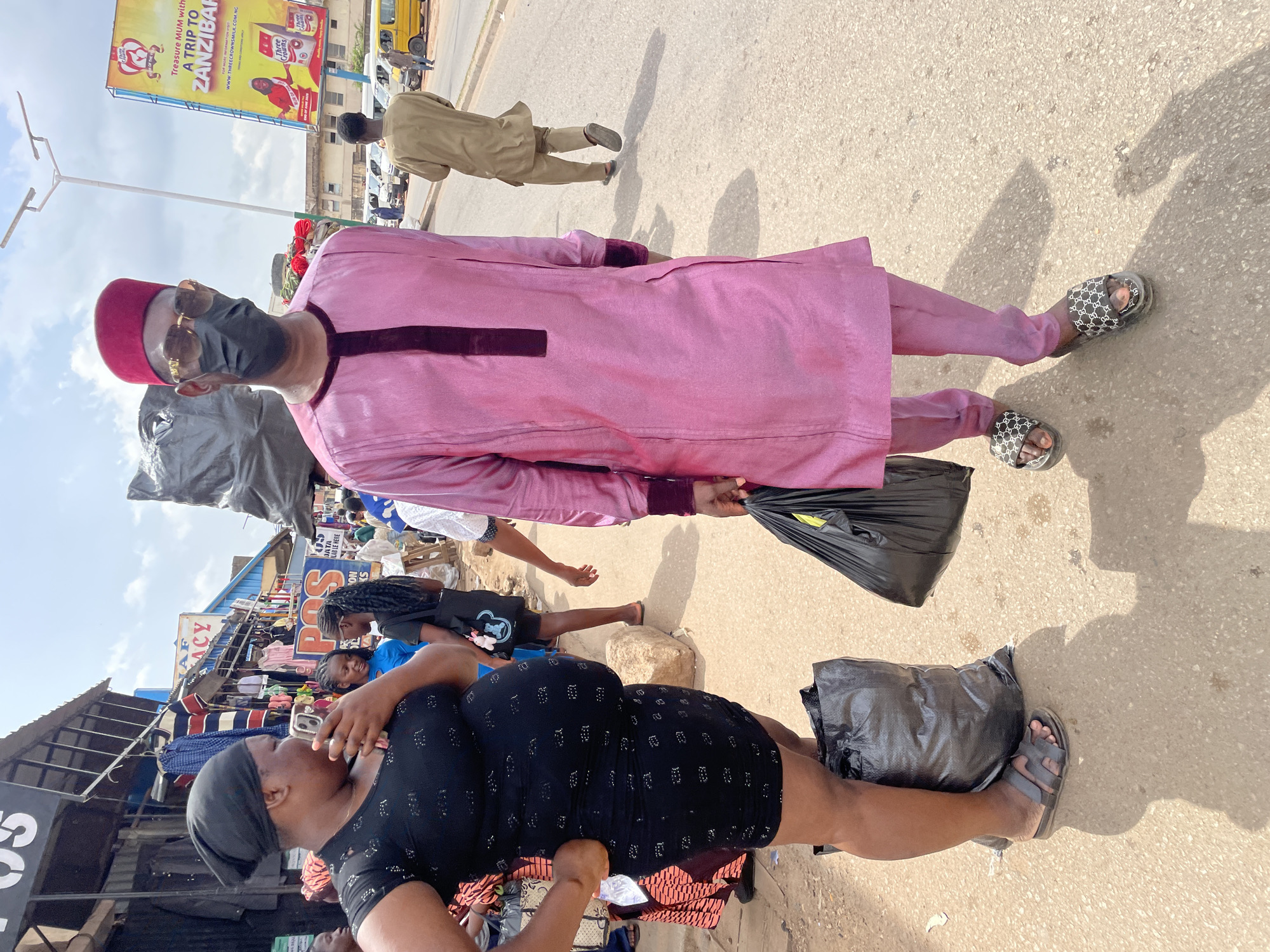 The image is a street scene in Jos, Nigeria. A man wearing a pink outfit, red fez, sunglasses, and a face mask stands prominently on the right side of the frame, holding a black plastic bag. He is wearing patterned sandals. In the foreground on the lower left is a woman with a black head covering and a dark patterned dress. She appears to be using her phone. A black plastic bag is on the ground near her. In the background are market stalls and other people. A yellow bus and a billboard are visible above and behind the man, advertising "A TRIP TO ZANZIBAR." The street surface appears to be paved. Another man is walking across the street behind the pink-clad man, wearing tan clothing.