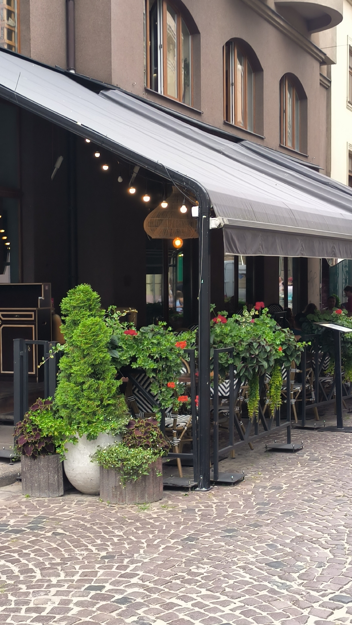 The image depicts an outdoor seating area of a cafe or restaurant in Lviv, Ukraine, situated on a cobblestone street. A dark brown building with arched windows forms the backdrop. The seating area is covered by a grey awning and features several tables with chairs, some with black and white patterned cushions. Lush greenery, including potted plants and flowering bushes with red and orange blossoms, adorns the front of the seating area. String lights hang under the awning, and a large woven light fixture is suspended from the ceiling. In the foreground, there are large concrete planters containing more plants. The pavement is made of irregular-shaped cobblestones. The overall scene suggests a pleasant outdoor dining or relaxing space.