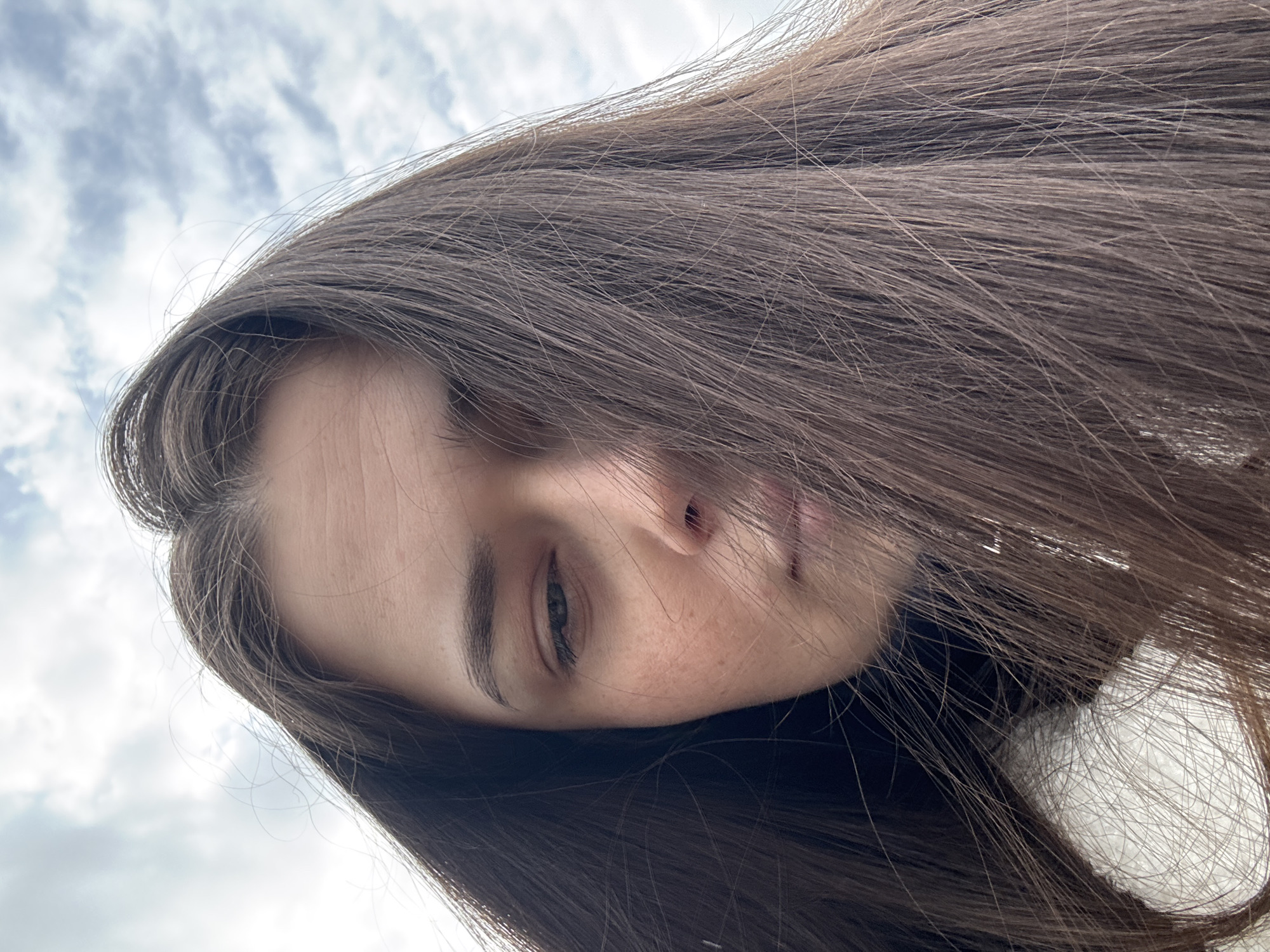 An extreme close-up, low-angle shot depicts a young woman's face partially obscured by her long, brown hair. The sky, filled with white and gray clouds, is visible in the background. The woman has fair skin, with visible freckles near her nose. Her dark eyebrow and eye are partially visible. The image's location, based on the prompt, is Chaikovsky, Russia, but this cannot be verified from the image alone.