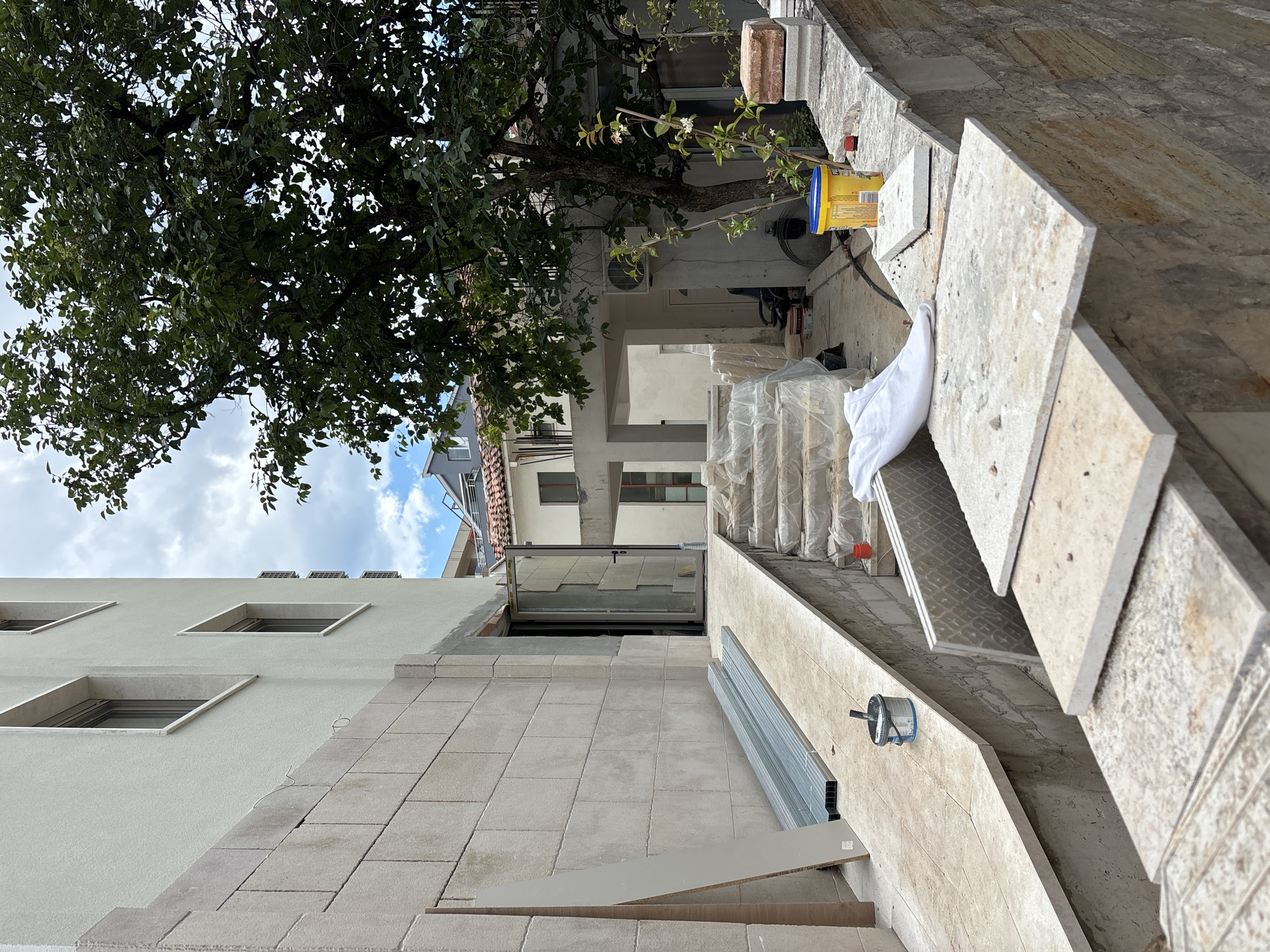 The image depicts an outdoor construction site located in Budva, Montenegro. A building under construction is visible on the left, with a light-colored stucco facade and a lower section clad in stone blocks. A ramp leads up to a doorway, and adjacent to it are a set of stairs covered in plastic sheeting. Construction materials, including metal pipes, tiles, and buckets, are scattered around the site. A large tree with lush green leaves is prominent on the right side of the frame, casting shadows. The sky is partially cloudy. The scene suggests ongoing renovation or new construction work in a semi-urban environment.