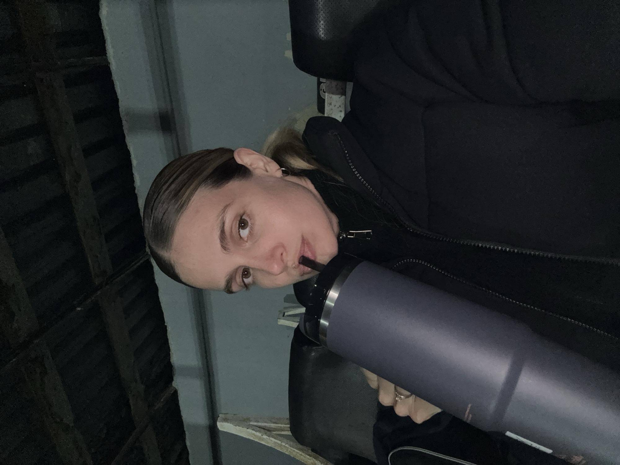 The image, rotated 90 degrees counter-clockwise, features a young woman looking directly at the viewer with a neutral expression. She is in a dimly lit, possibly indoor or covered setting, suggesting night or low-light conditions.
The woman has light brown hair, pulled back, and is wearing a black zippered jacket. A small hoop earring is visible in her left ear. She holds a large, dark grey or purple-blue tumbler with a silver rim in her right hand, from which she is drinking through a black straw. A silver-toned ring is visible on her right hand.
The background on the left reveals dark, exposed wooden beams or rafters, indicative of a workshop, garage, or industrial-style ceiling. To her right, a roughly textured, light grey wall is visible. The environment appears functional and unadorned. No visible text is present in the image. The location context for this scene is Tbilisi, Georgia.