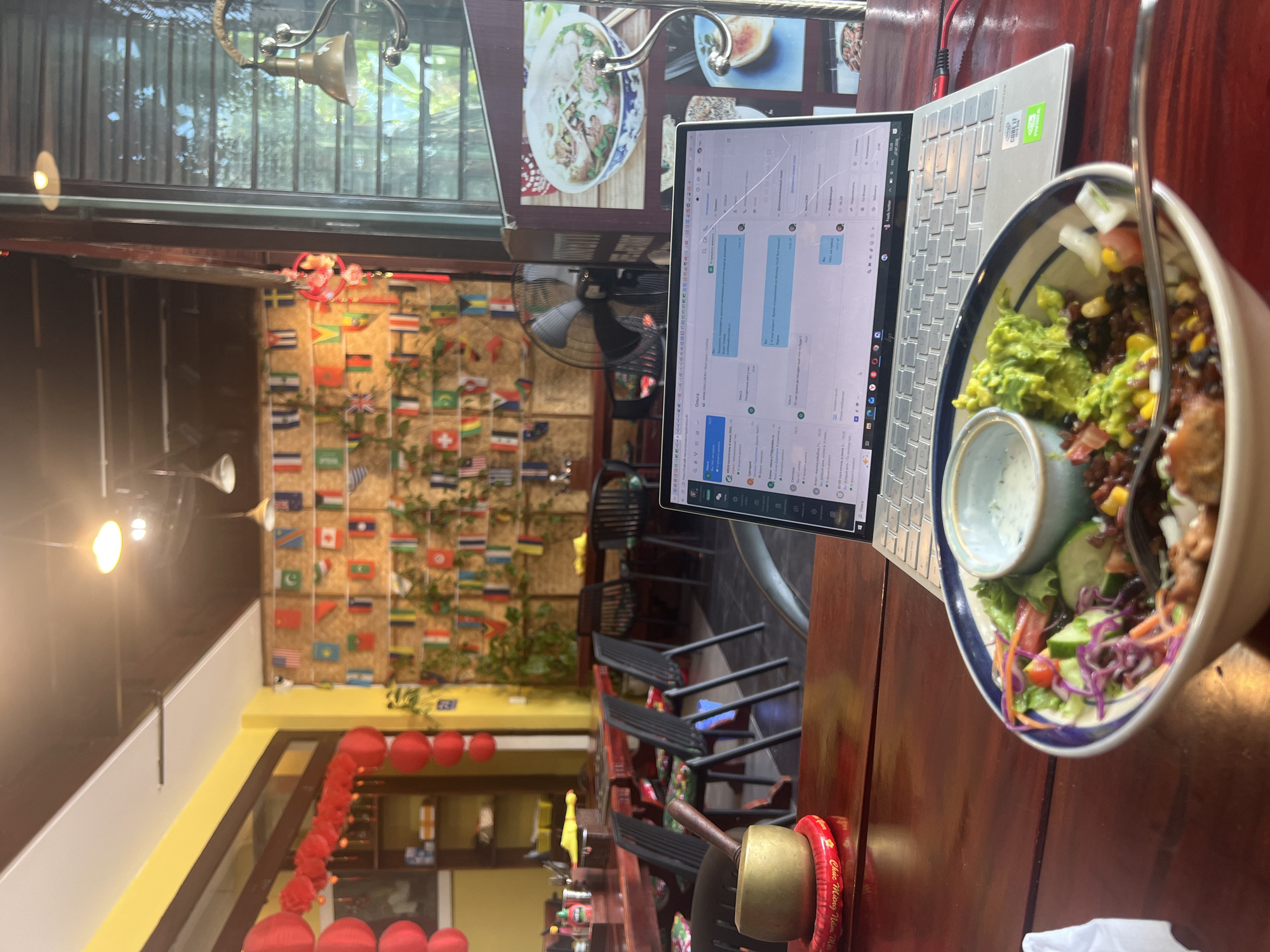 The image depicts an indoor scene at what appears to be a restaurant or cafe in Hoi An, Vietnam. In the foreground, a wooden table holds a laptop displaying a messaging application and a bowl of food that includes salad, avocado, and beans. To the left, a brass singing bowl rests on a red circular mat with Vietnamese writing. The background features a wall decorated with numerous small national flags, suggesting an international theme. Red lanterns are visible on the left side, and beyond them, tables and chairs are arranged, indicating a dining area. A large fan is positioned further back. The overall ambiance suggests a casual dining and working environment.