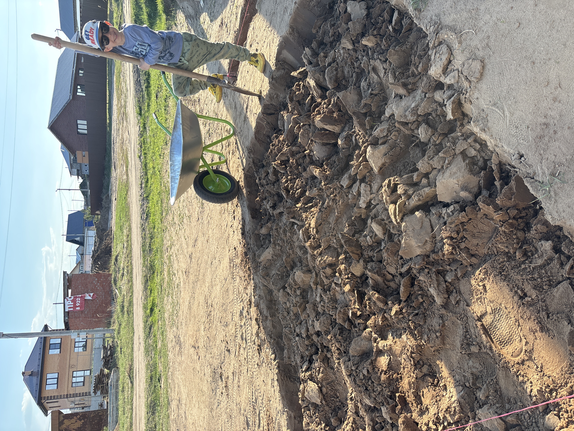 A child, wearing sunglasses and a cap, stands next to a green wheelbarrow, holding a shovel as if leaning on it. The child is working in a large dirt area. Behind the child, there are various buildings, with one appearing to be under construction or recently completed, displaying a sign in Russian ("ПРО 8 932 3"). The location appears to be in Chaikovsky, Russia, based on the sign. The scene appears to be a construction or landscaping project.