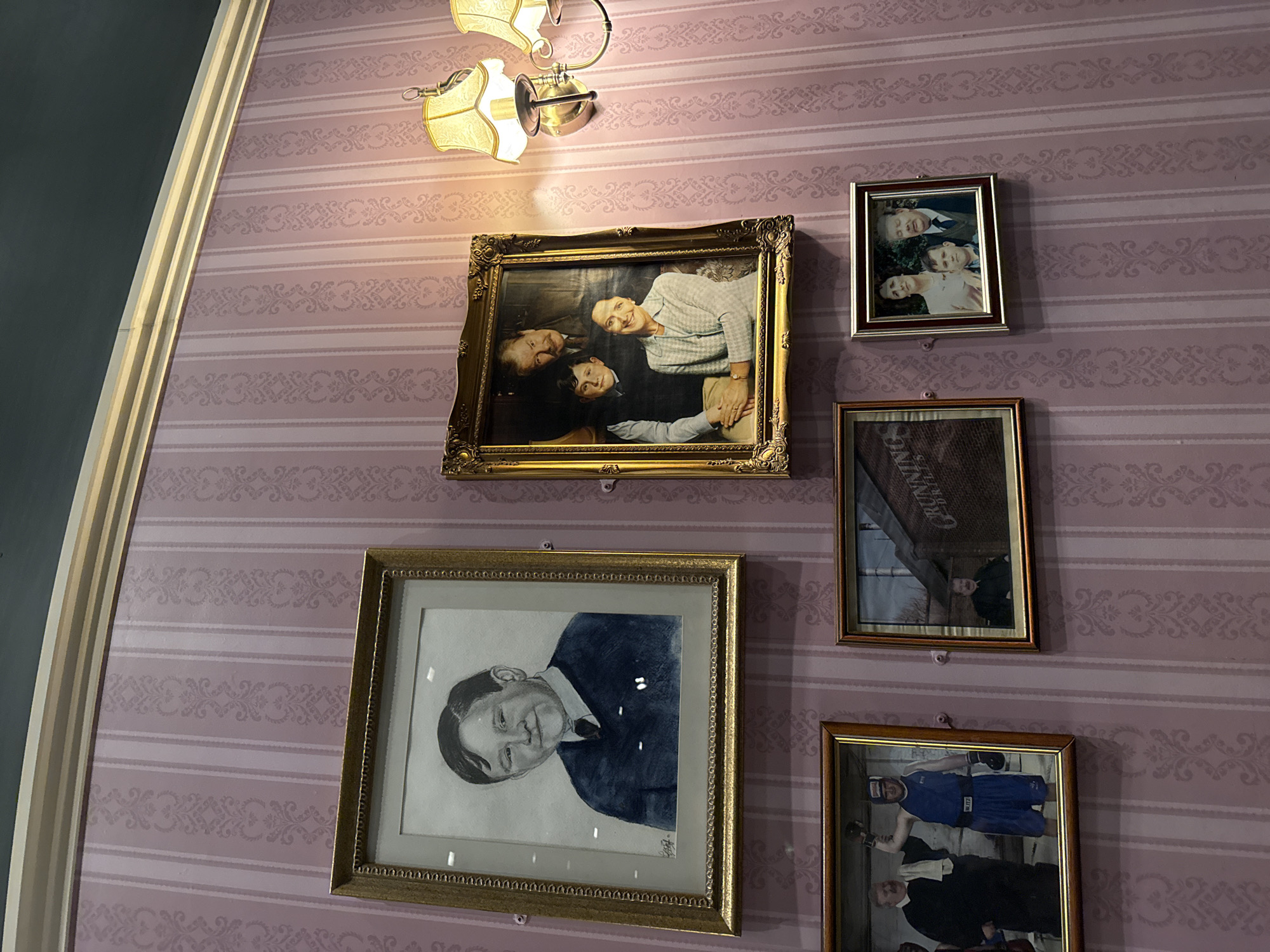 The image shows a collection of framed photographs and portraits hanging on a wall with pink striped wallpaper adorned with a repeating damask pattern. The wall is lit by two ornate wall sconces with yellow lampshades.
In the upper left is a framed black and white portrait of a young boy in a dark sweater and tie, his hair parted to the side. To the right of this, a larger, ornate gold-framed portrait depicts a family of three: a man with a mustache and dark suit stands behind a young boy in a sweater vest, while a smiling woman in a patterned jacket sits in the foreground.
Below the first portrait, on the left, is a framed photograph of two men, one in a priest's robe and the other in a blue boxing outfit with gloves raised in victory. To the right of this is a framed photograph showing a man sitting in front of a brick building with the word "GRUNNING" visible. Further to the right and slightly lower is a smaller framed photograph of a family of three, including a young boy in the center, with a man and woman standing behind him.
The image appears to be taken indoors, possibly in a public space or a home with a decorative aesthetic. The location is identified as Three Rivers, United Kingdom, though no specific landmarks or context beyond the decorative elements and photographs are provided.