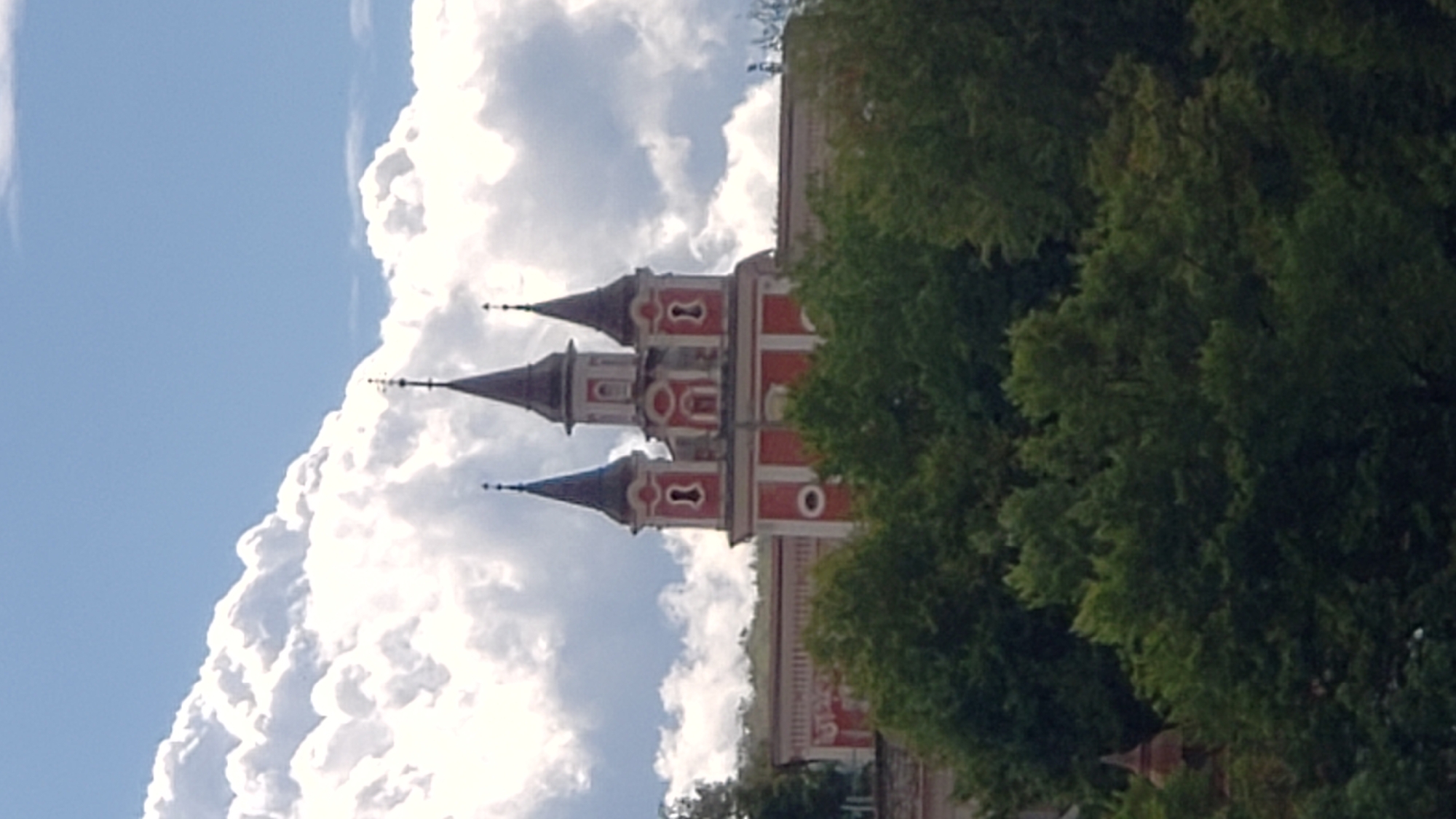 A photograph captures a section of the Church of the Holy Spirit in Prešov, Slovakia, set against a backdrop of a bright blue sky with dramatic white clouds. The church features a prominent red facade with white architectural details, including arched windows and decorative elements. Three dark spires rise above the main structure, each topped with a cross or spire-like ornament. The building is partially obscured by lush green trees in the foreground, which extend across the lower portion of the image, suggesting the church is situated on elevated ground or surrounded by dense foliage. The overall impression is one of historical architecture set within a natural landscape.