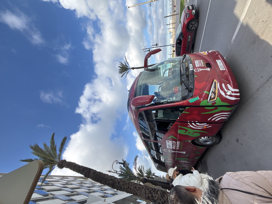 A large red Irizar touring coach, branded for the "CAF Africa Cup of Nations 2023" with green and red Moroccan-themed patterns, is stationary or moving slowly on an asphalt road in Tangier, Morocco. The bus displays a Moroccan license plate ending in '11', which designates Tangier. Several individuals are gathered on the sidewalk to the left of the bus; one person in a white jacket and baseball cap is observed looking at a mobile device. A red crossover SUV is present on the road to the right of the bus, moving away from the viewer. The scene is set under a blue sky with scattered white clouds. A tall palm tree stands prominently on the left, next to a modern multi-story building. Other palm trees and streetlights are visible. In the background, beyond the road and vehicles, a maritime environment consistent with a port or marina is discernible.