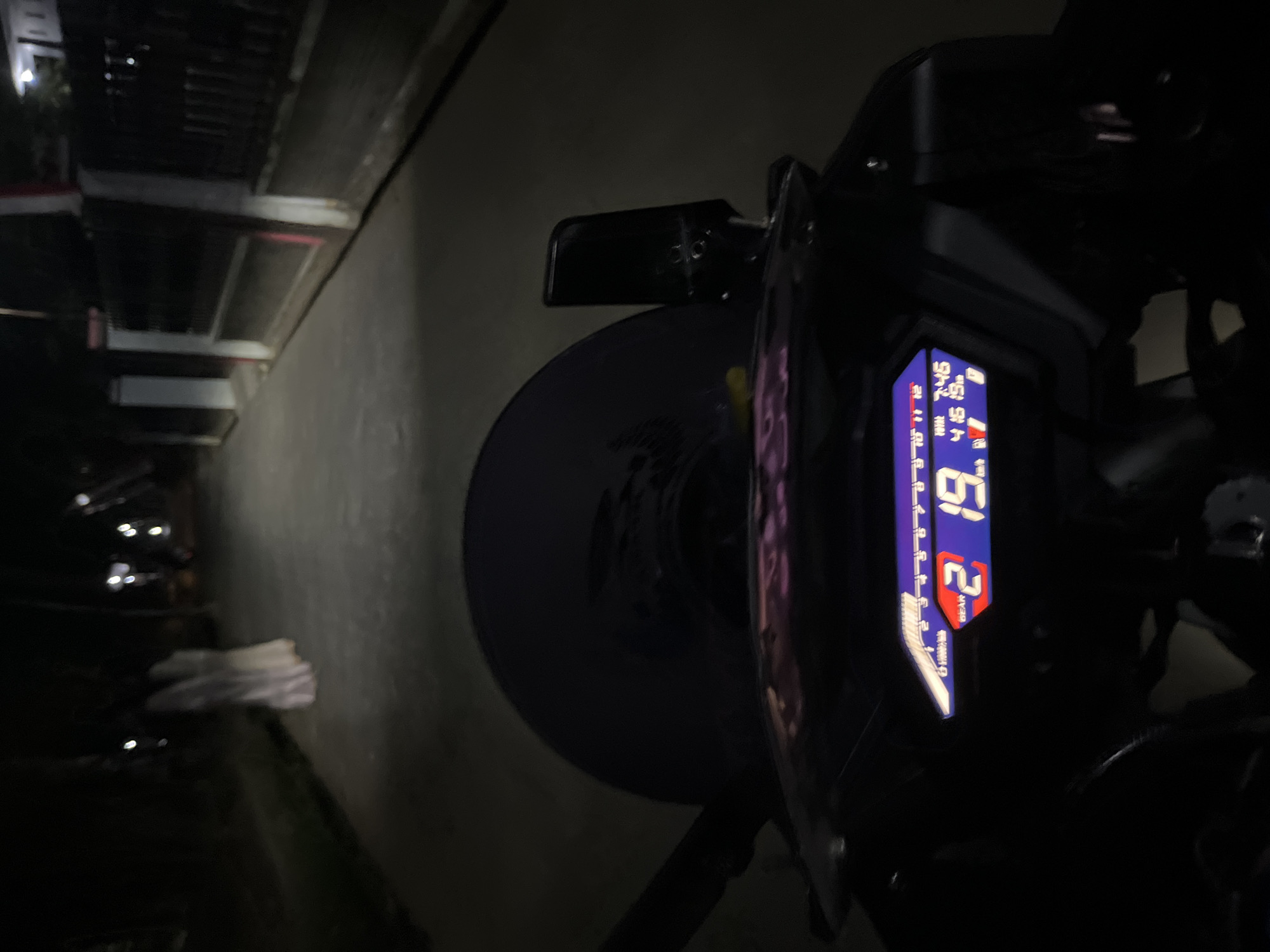 The image is a first-person perspective from a motorcycle at night, likely in Karangrandu, Indonesia. The motorcycle's digital dashboard is illuminated, showing the gear is "2", the speed is "19" (presumably km/h), the time is "7:45", and the date is "4/16/19". The road ahead is paved and dimly lit by the motorcycle's headlight and distant streetlights. On the left side of the road, a person is visible in the distance, wearing a long, white garment, appearing to be walking. On the right side of the road, there is a fence with vertical posts and horizontal bars, and a building is visible beyond it. The overall environment suggests a quiet, possibly residential street at night.