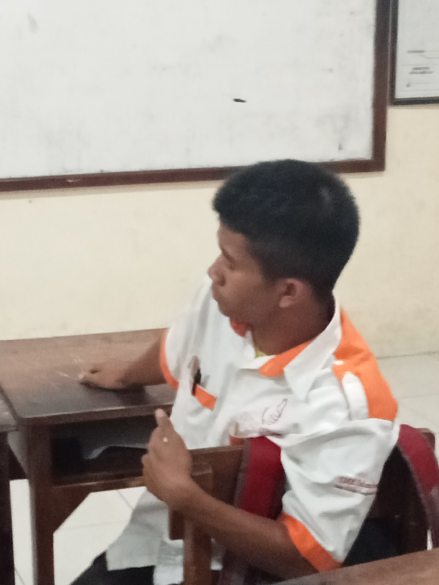 The image shows a young person sitting at a wooden desk in what appears to be a classroom. The person is wearing a white polo shirt with orange accents on the collar and sleeves. Their head is turned to the right, away from the camera, and their mouth is slightly open. There is a whiteboard behind the person, and the walls appear to be a light color. The desk has a storage compartment underneath. The overall scene suggests a casual setting, possibly during a lesson or break. The specific location in Secang, Indonesia, cannot be determined from the image alone.