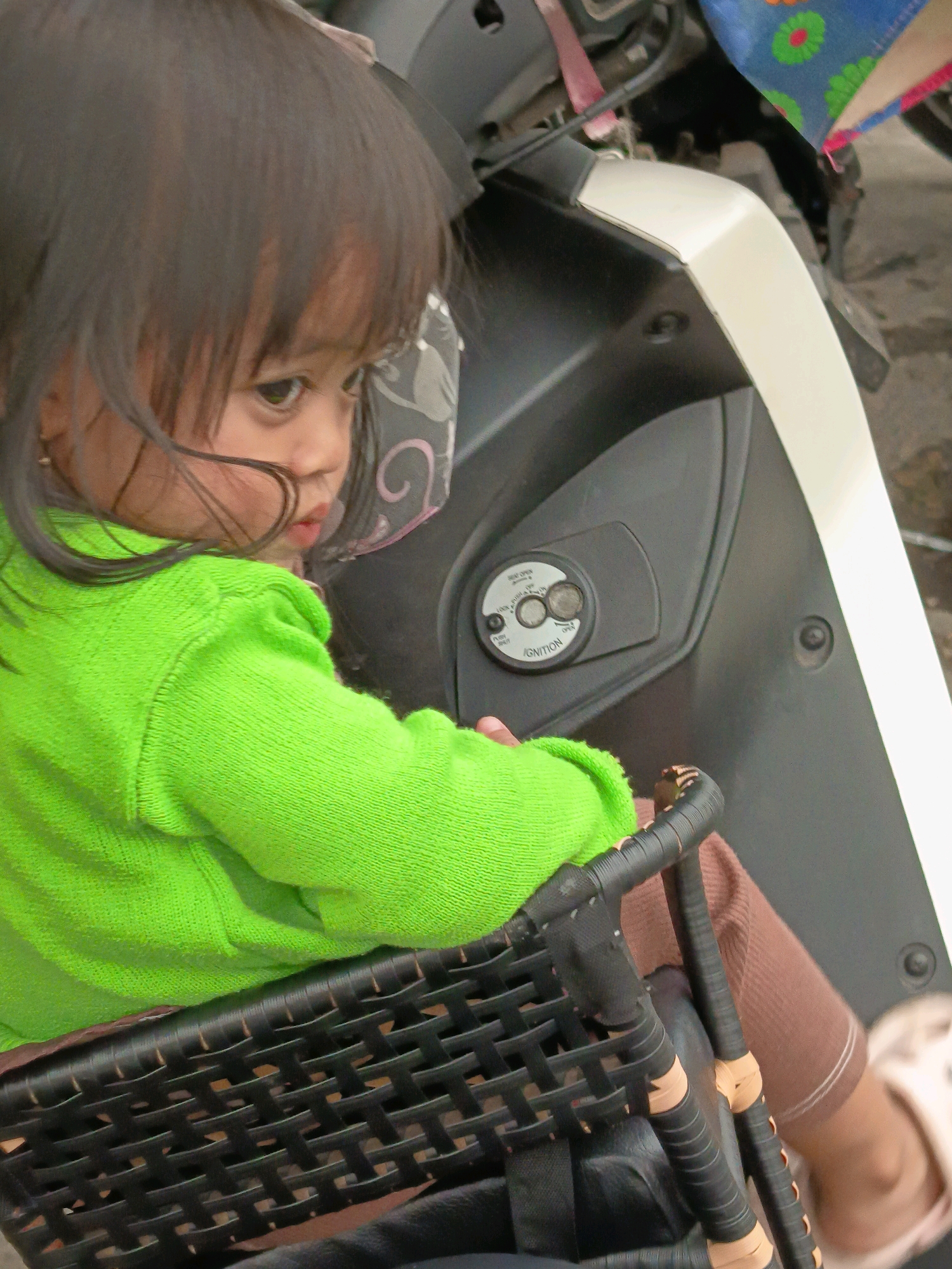 The image shows a close-up of a young child, possibly a girl due to her dark hair and earrings, sitting in a black woven seat. She is wearing a bright green knitted sweater. The child is facing away from the camera, looking to the right. Her dark hair is falling over her face and shoulders. Her right arm is bent and resting on the seat.
Behind the child, the dark grey and white body of a scooter is visible. On the grey body, near the center of the image, is the ignition switch of the scooter. The switch has several positions labeled "LOCK", "OFF", "ON", and "OPEN". A blue and multi-colored fabric item, possibly a flag or a cloth, is draped over the handlebars in the upper right portion of the image. The background appears to be an outdoor setting, with a slightly out-of-focus ground that could be pavement or dirt. The overall lighting suggests it is daytime.
The provided context places this scene in Banjarmasin, Indonesia.