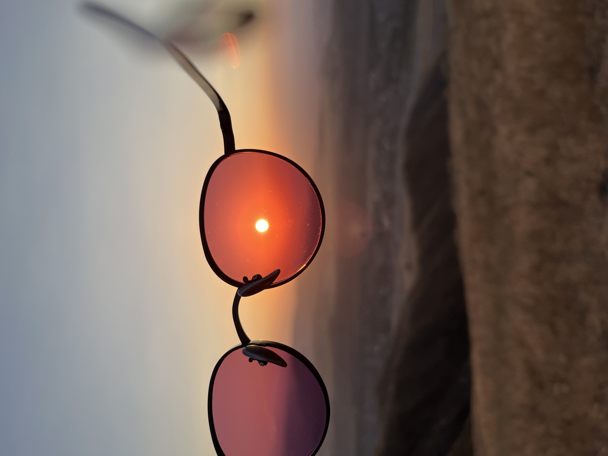 A close-up shot focuses on a pair of dark-rimmed sunglasses held up to the camera. The lenses are tinted with a warm, reddish-orange hue, reflecting the setting sun which appears as a bright, circular orb within the right lens. The background, seen through the lenses and beyond, depicts a hazy, mountainous landscape with rolling hills and sparse vegetation, indicative of an outdoor, possibly rural or natural setting, such as the outskirts of Besh Kungei, Kyrgyzstan. The sky above transitions from a soft blue at the top to a gradient of orange and pink near the horizon, suggesting dusk or dawn. The foreground consists of dry, golden grass, adding to the natural, open-air feel of the scene.
