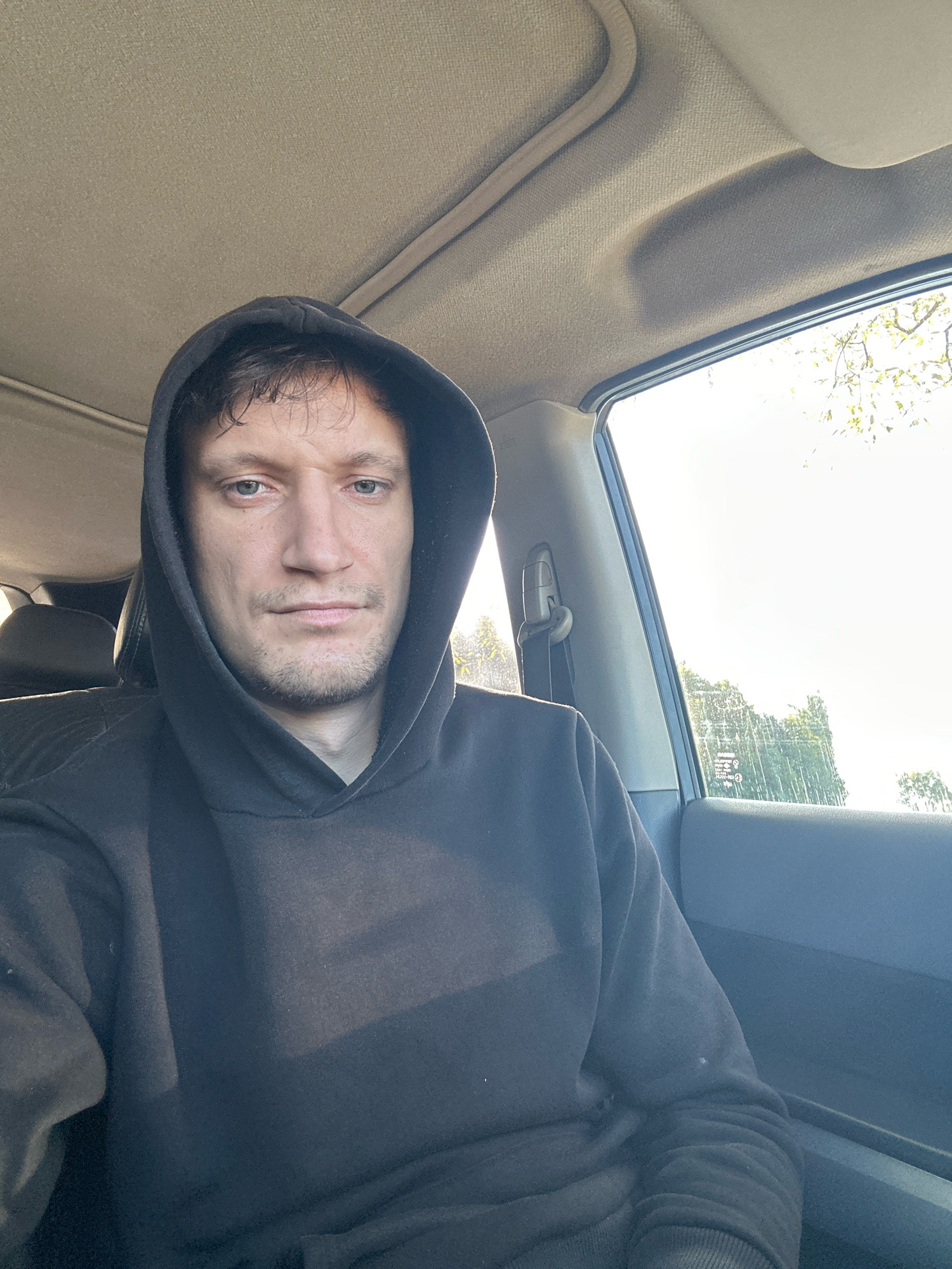A man wearing a dark hoodie sits in the passenger seat of a vehicle. The man's face is partially obscured by the hood. He has light skin, brown hair, and light-colored eyes. He is looking directly at the camera with a neutral expression. The interior of the car is visible, including the headliner, a sunroof, and the passenger-side window. Through the window, trees and a bright sky are visible. There are no discernable landmarks or indicators of the specific location within Makiv, Ukraine.