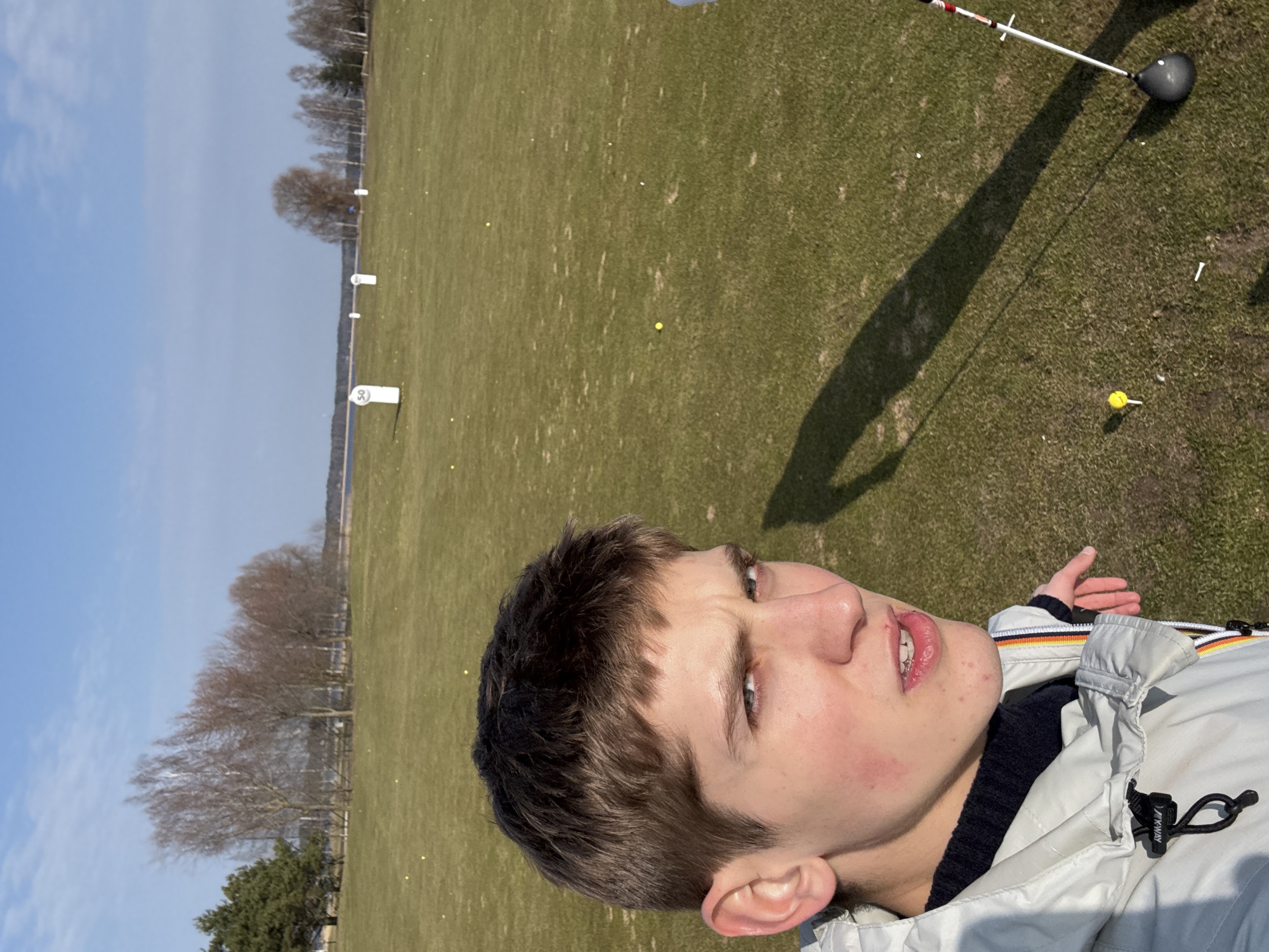 The image depicts a fair-skinned young man in a white K-Way jacket with a black knit sweater underneath. He has short brown hair and is looking upwards and slightly to the left. He appears to be on a golf driving range. In the background, there is a large expanse of grass with distance markers at 50, 100, and further away. A golf club is visible in the upper right corner, casting a shadow on the grass. The sky is blue with scattered clouds. Based on the vegetation and landscape features, the location is likely in or near Riga, Latvia. The objects of focus in the photograph are a golf club, a person, a golf range, and the clear sky. The action seems to be the person at the golf range.