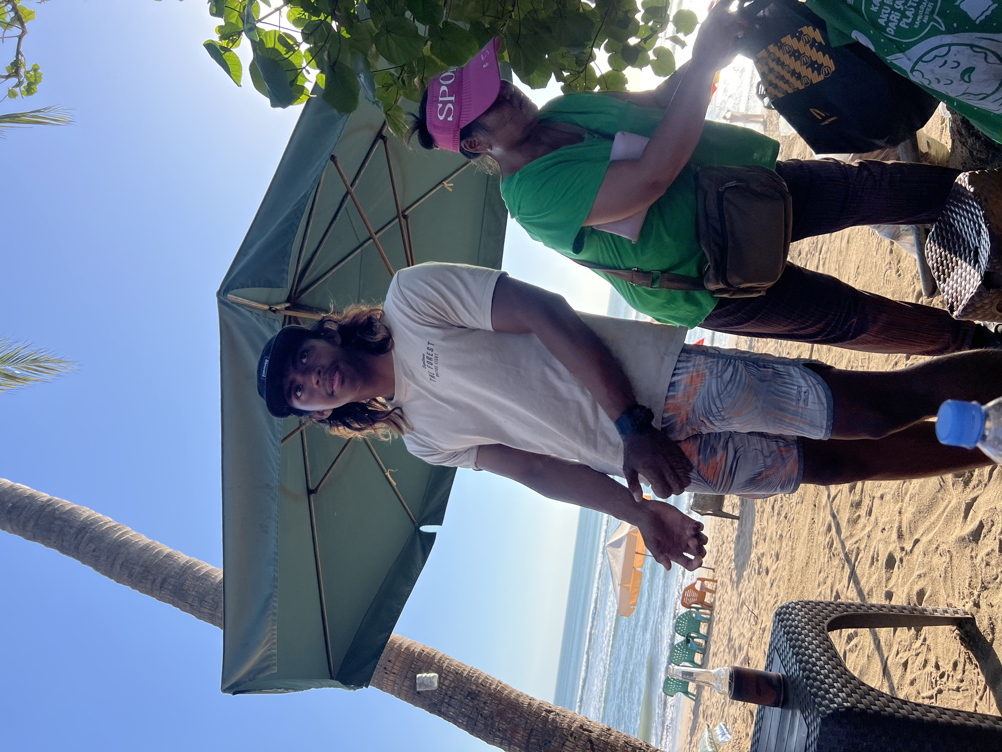 The image depicts a beach scene in Kuta, Indonesia, likely near a beachfront cafe or bar. In the foreground, a young man with long, dark hair and a baseball cap stands under a large green umbrella, wearing a light-colored t-shirt and patterned shorts. He has his hands outstretched, palms up, as if gesturing or asking for something. To his right, a woman wearing a pink visor and a green t-shirt is bent over, rummaging through a bag. A dark brown crossbody bag is slung over her shoulder.
In the immediate vicinity, a black woven table holds a glass bottle of dark liquid and is placed on the sandy beach. Further back, several colorful plastic chairs are scattered on the sand, facing the ocean. The ocean itself is visible in the background, with gentle waves rolling towards the shore. A tall palm tree stands to the left of the frame, its trunk partially visible. The overall lighting suggests it is either early morning or late afternoon, with the sun creating bright highlights and casting shadows. The environment is characterized by sand, the sea, and beachside amenities.