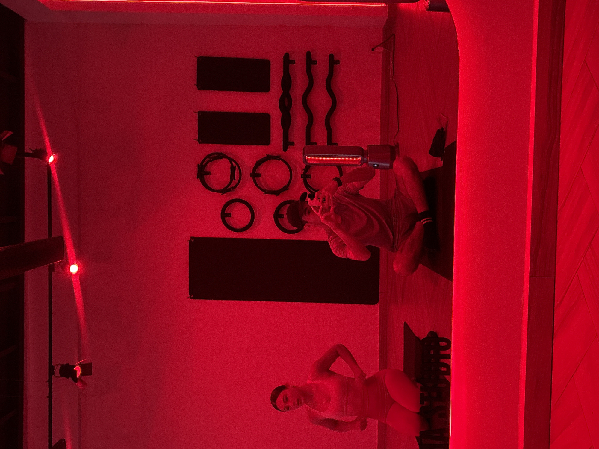 A brightly lit, red-hued interior appears to be a workout space. Gym equipment like circular bands, curved bars, and rectangular mats adorn the walls. Two people are present: One person is seated, and another woman is standing, with one hand on her waist, wearing what appears to be workout attire. The location is likely San Bernardino Tlaxcalancingo, Mexico.