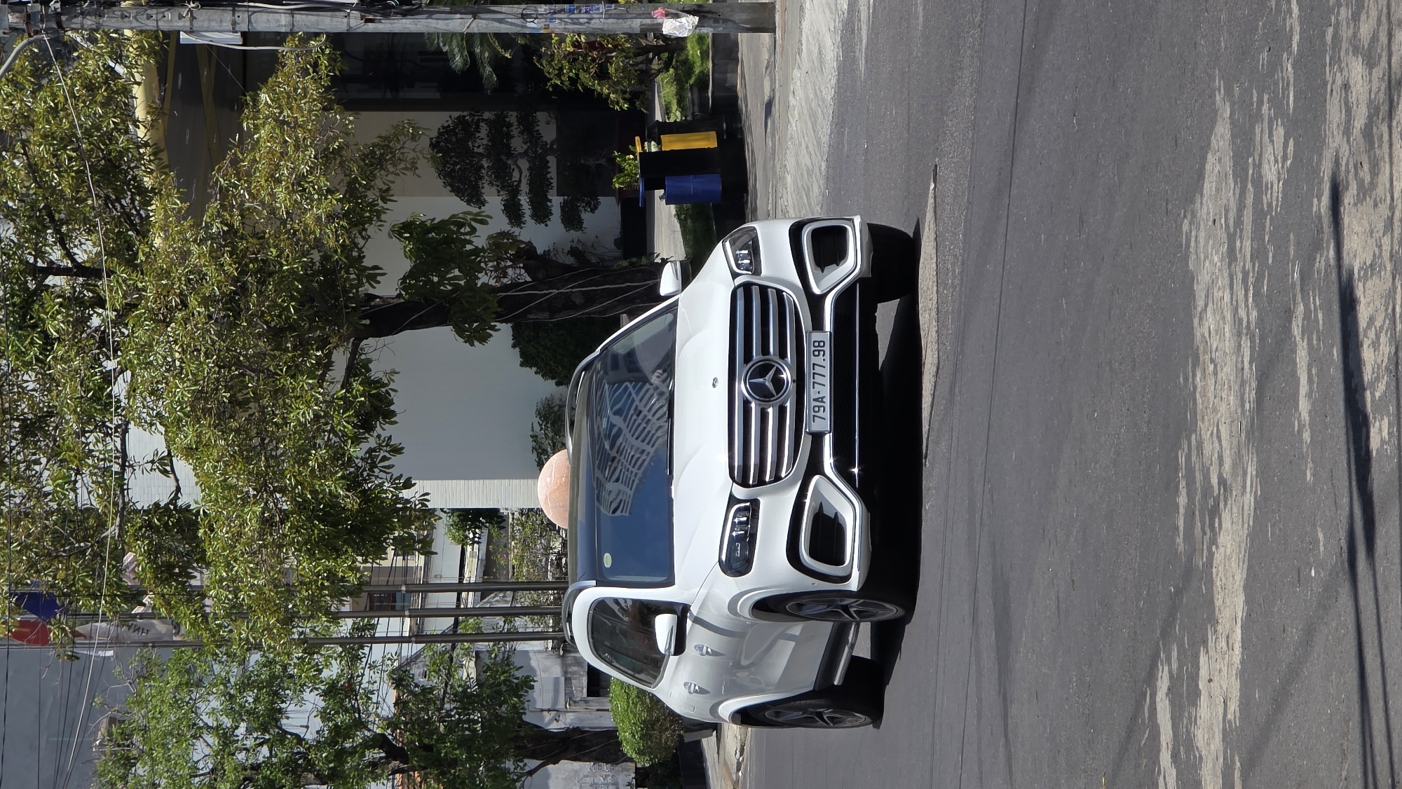 A white Mercedes-Benz GLC is parked on an asphalt road in Bắc Nha Trang, Vietnam. The front of the car faces the viewer. The license plate reads "79A-777.98." The car is parked near a tree and a building. Shadows from trees are visible on the road. Overhead power lines are also present.