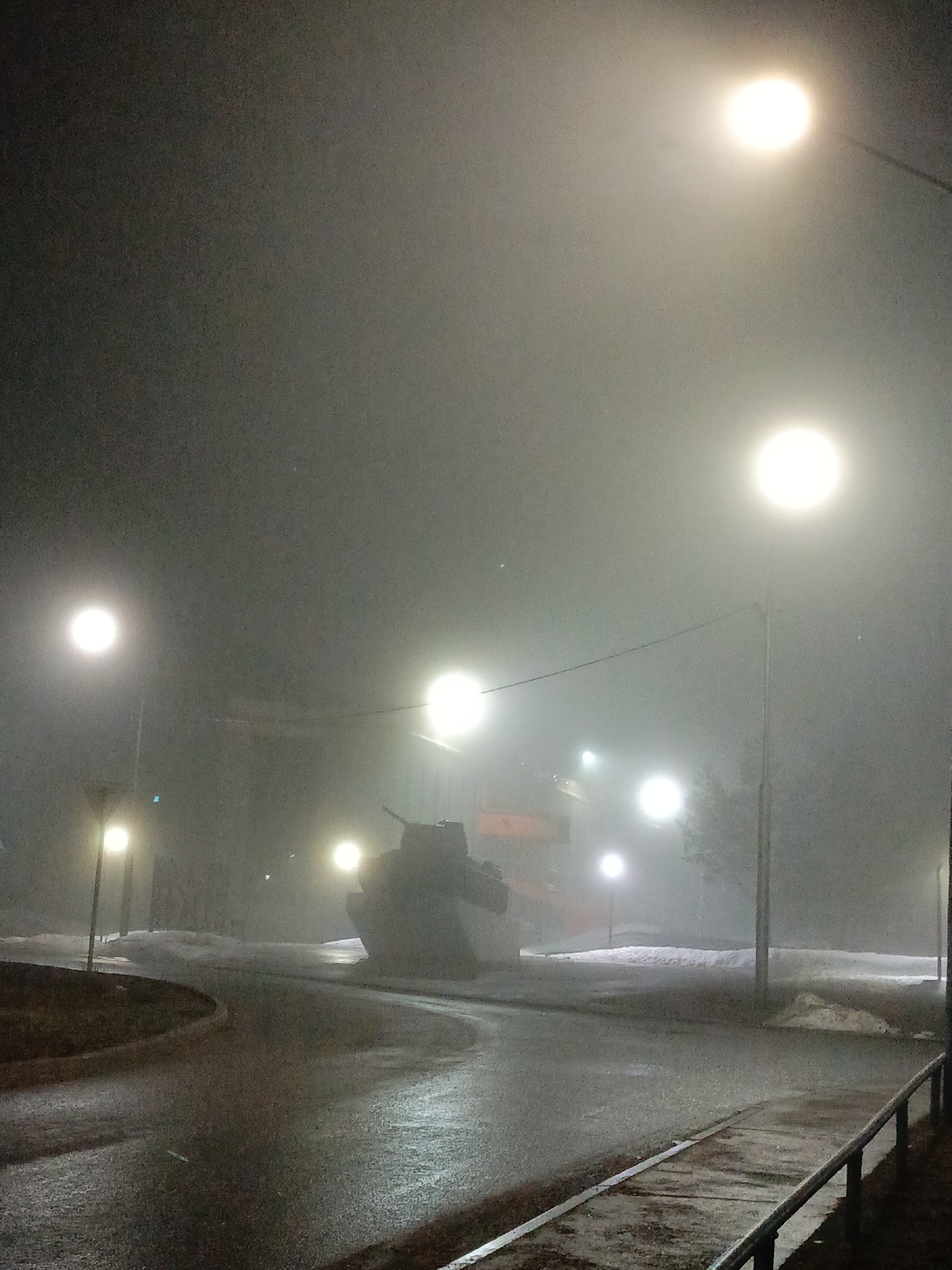 The image shows a foggy night scene in Марковский, Russia. The foreground features a wet asphalt road curving around a traffic island with grass. A sidewalk with a railing is visible on the right side of the image. Several streetlights illuminate the scene, creating a bright glow in the fog. A large, dark object resembling a tank or armored vehicle is positioned in the center of the scene, partially obscured by the fog. Snow patches can be seen on the ground near the road. In the background, more streetlights and some blurry structures can be distinguished through the fog. The overall impression is a cold, damp, and somewhat eerie atmosphere.