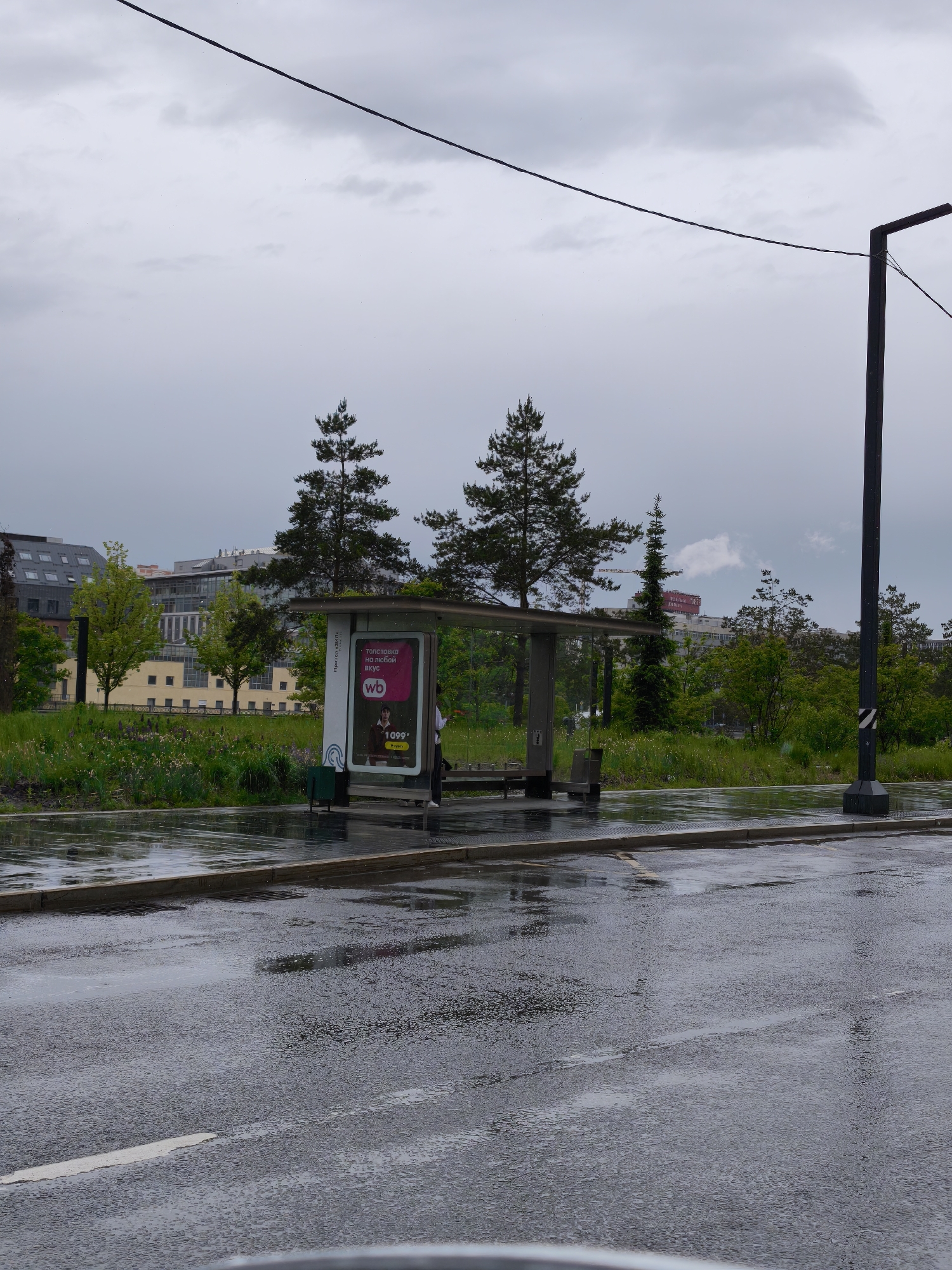 The image shows a wet street in Moscow, Russia. A bus stop is visible on the opposite side of the road. The bus stop has an advertisement for "wb" (Wildberries), showcasing a sweatshirt for 1099 rubles. A person is standing near the bus stop, possibly waiting for a bus. In the background, there are trees and buildings, including a building with a red sign on top. The sky is overcast, indicating rainy or cloudy weather. A lamppost stands near the bus stop. Power lines are visible overhead. The street is wet and reflects the surroundings.
