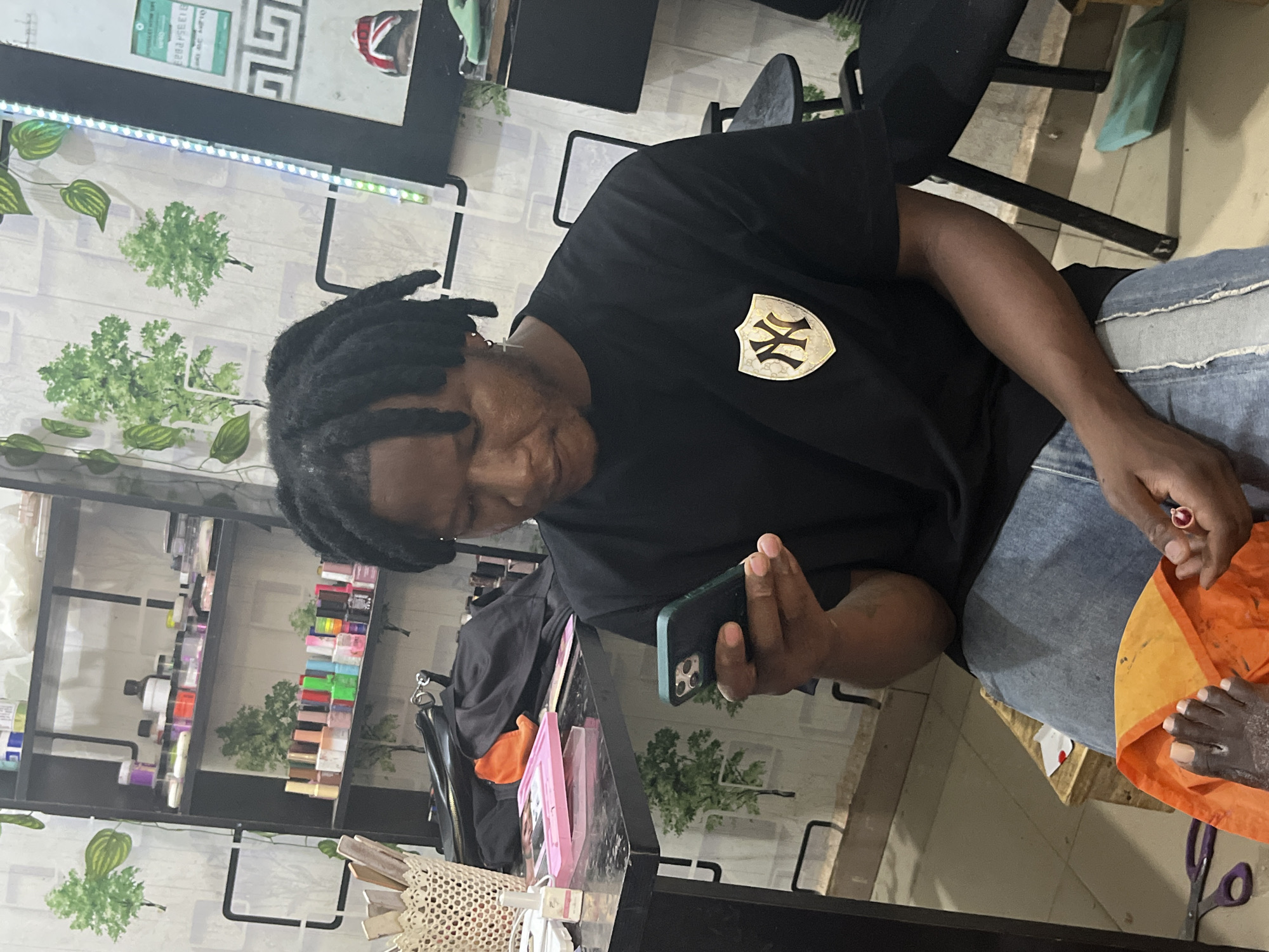 An adult male with dreadlocks is seated, observing a dark-colored smartphone held in his left hand. He wears a black short-sleeved t-shirt featuring a gold and white New York Yankees logo on a shield emblem on the left chest, and distressed blue denim jeans. A small cross earring and hoop are visible in his right ear. His bare left foot rests on an orange fabric draped over a wooden stool, indicating a pedicure is either in progress or awaiting service. His right hand holds a small red cylindrical object, possibly a nail tool.
The immediate foreground features a black table holding a white wicker basket containing wooden sticks, a small white bottle, and a pink rectangular tray. A black handbag and an orange piece of clothing are also on the table. On the floor beside him are purple-handled scissors and a small white packet with a red mark.
The background shows a black shelving unit against a white wall decorated with green tree and leaf wallpaper motifs. The shelves are stocked with numerous small containers of various colors, consistent with nail polishes, gels, and powders found in a beauty salon or nail parlor. A vertical LED strip light and a mirror reflecting a shoe with a Union Jack flag design are visible on the right wall. The floor is tiled.
The specific location within the city of Jos, Nigeria, cannot be determined from the visual information present in the image.