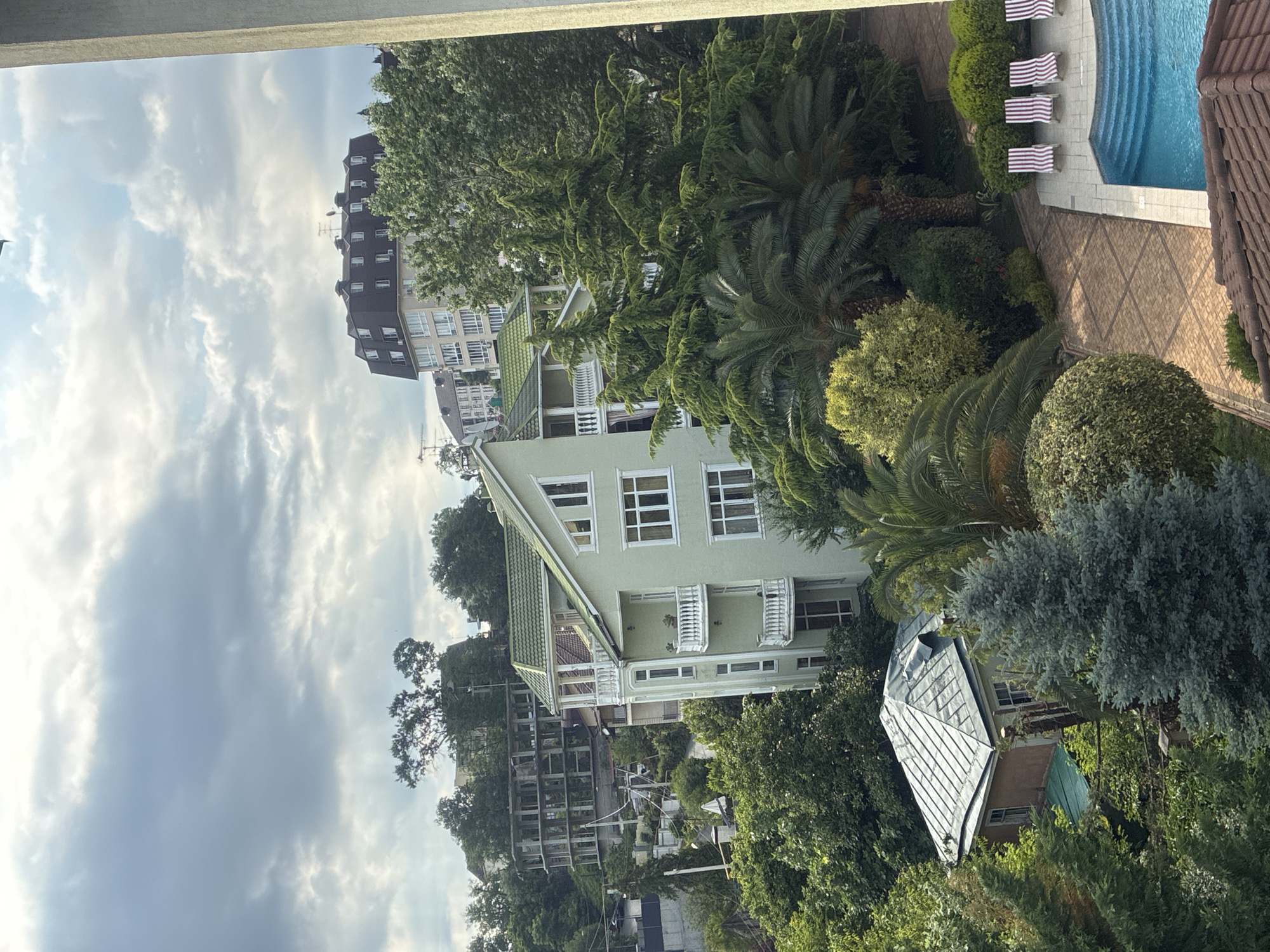 This is an aerial view of a resort area in Sochi, Russia, featuring a light green multi-story building with white balconies and a green roof, surrounded by lush trees and palm trees. In the foreground, a small, quaint house with a metallic roof is nestled amongst the foliage. To the right, a swimming pool with blue water and tiered steps is visible, accompanied by a paved area with several lounge chairs. Further in the background, other buildings, including a large residential complex with a dark mansard roof, are situated on a hillside under a cloudy sky. The scene appears to be a tranquil, residential or hotel complex.