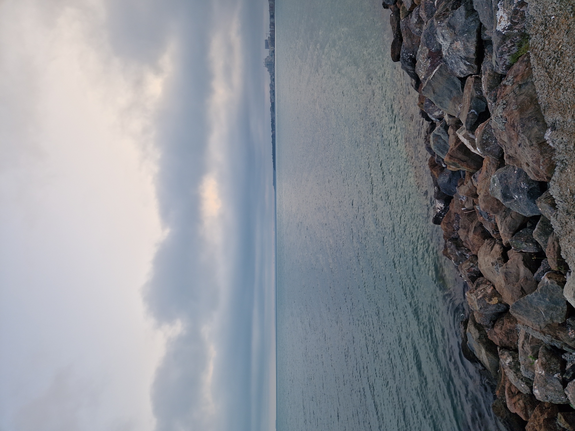 The image displays a wide panoramic view of a large body of water under a heavily overcast sky, with a rocky shoreline in the immediate foreground and a distant urban landscape on the horizon.
In the foreground, a substantial array of dark grey, brown, and reddish-brown rocks forms the edge of the land, partially submerged by the water. These rocks vary in size and shape, creating an uneven, natural barrier. A small amount of light-colored gravel and dirt is visible at the very bottom right, adjacent to the rocks.
The midground is dominated by a vast expanse of calm, dark teal-blue water. Gentle ripples are present across its surface, suggesting minimal wave activity.
On the distant horizon, a strip of land hosts a continuous line of light-colored, multi-story buildings, indicating a developed city or coastal town. These structures appear somewhat indistinct due to the distance and atmospheric conditions.
The sky is filled with thick, grey clouds, stretching from one side of the frame to the other. Patches of lighter, softer illumination are visible within the cloud cover, particularly towards the upper middle and right, suggesting an indirect light source, possibly obscured by the clouds. The overall lighting is subdued and even, creating a tranquil and somewhat muted atmosphere. No individuals or specific activities are observable in the scene.