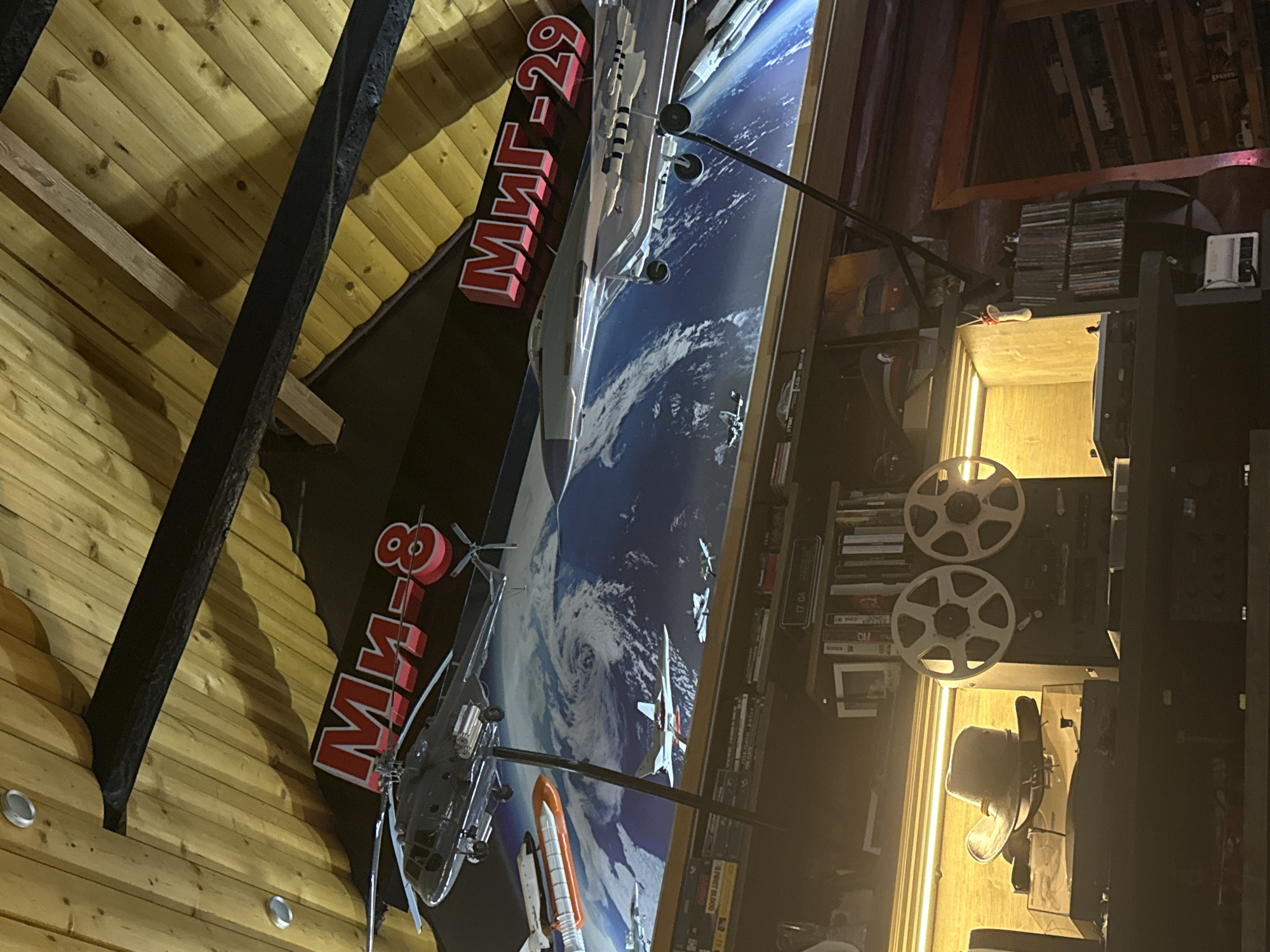 The image depicts an indoor scene with a wooden ceiling. In the upper portion of the image, there are prominent red Russian text labels for "Ми-8" (Mi-8) and "Миг-29" (MiG-29) displayed above a large mural or wallpaper that shows Earth as seen from space, with various model aircraft suspended in front of it. These models include a helicopter (likely the Mi-8) and a jet fighter (likely the MiG-29), as well as a space shuttle and other smaller aircraft.
Below the mural, a shelf is visible containing numerous items. On the left side of the shelf, a vintage reel-to-reel tape recorder is present, topped with a cowboy hat. To the right of the tape recorder are two large, white film reels. Further to the right, more shelves are filled with what appear to be books, CDs, or other media. The lighting in this lower section is warm and artificial, creating a cozy atmosphere. The overall impression is of a personal collection or display area, possibly in a home or a private hobby space. The location of Malaya Ven'ya, Russia is mentioned in the prompt but not visually evident within the image itself.