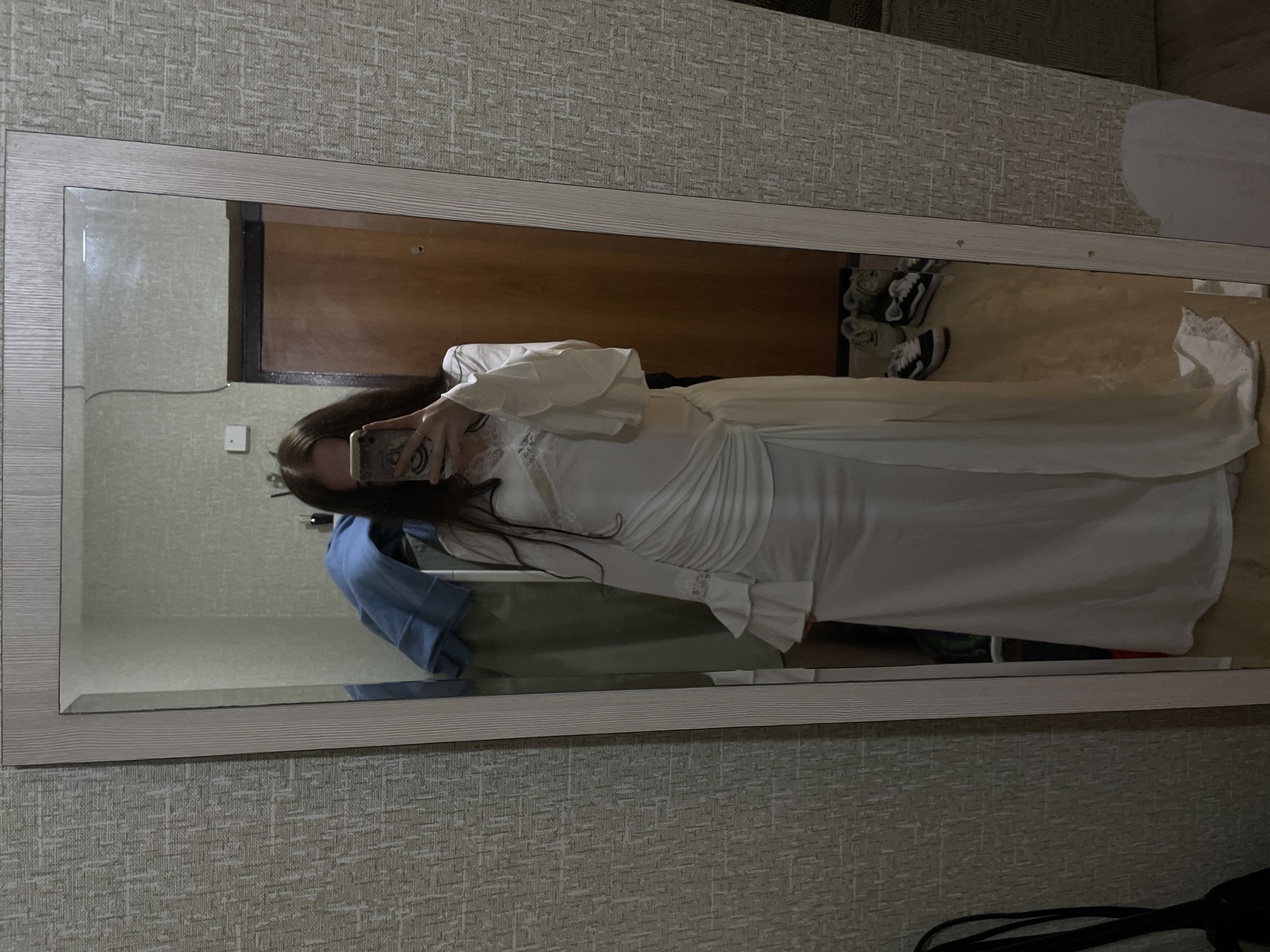 A young woman with long brown hair is standing in front of a full-length mirror and taking a selfie. She is wearing a flowing white dress with bell sleeves and lace detailing around the neckline. The dress is ruched and draped on one side, creating a cinched effect. Her hand holding a phone is visible in the mirror. In the reflection, a light-colored wall with a textured wallpaper pattern is visible, along with a wooden door and a closet with blue clothing. Several pairs of sneakers are on the floor in the reflection, near the bottom right of the mirror. The scene appears to be in a residential setting, likely a bedroom or dressing area, in Votkinsk, Russia.
