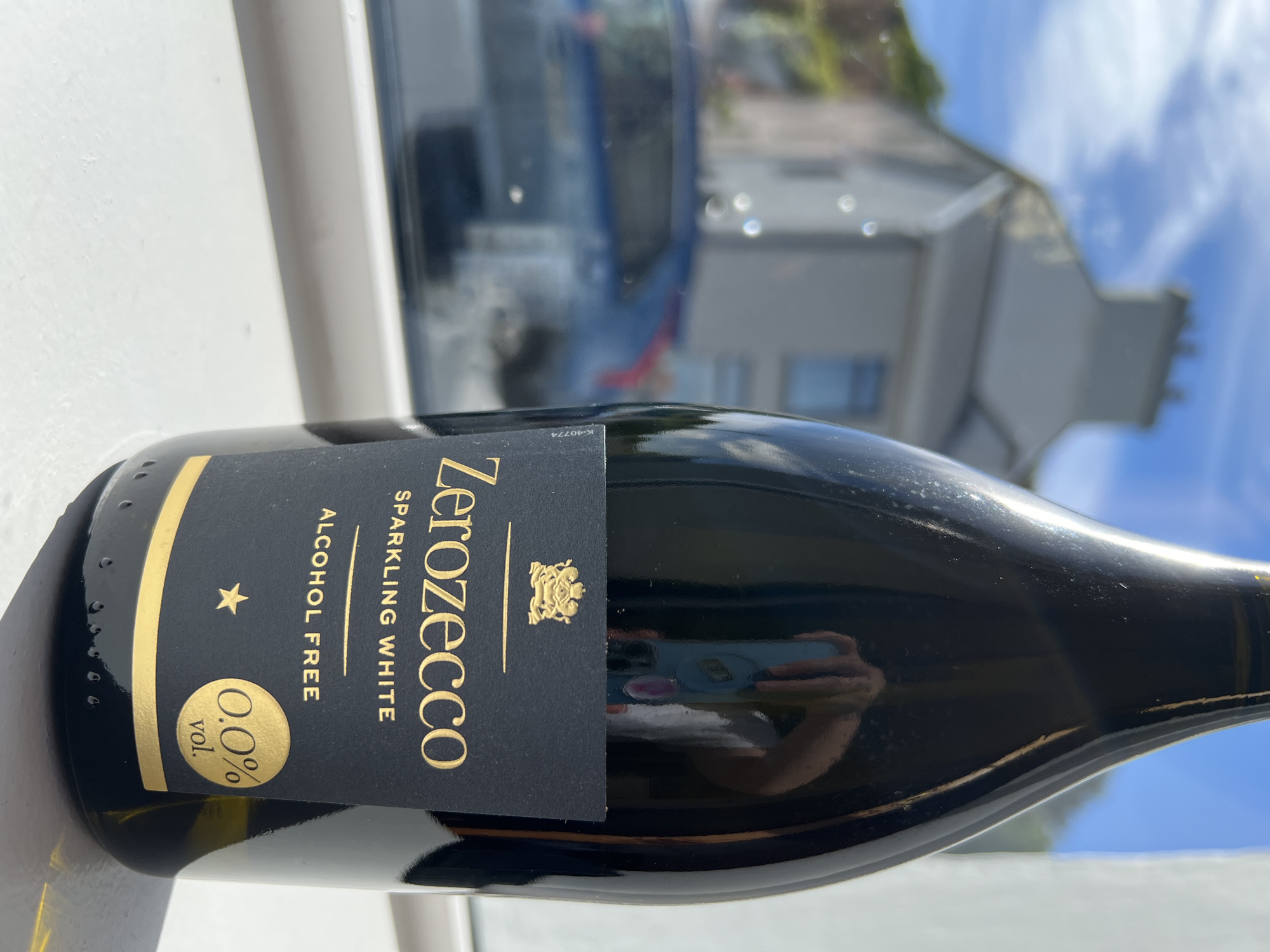 This media file features a close-up, slightly angled shot of a dark bottle of "Zerozecco" sparkling white, alcohol-free wine. The bottle has a prominent black label with gold lettering and accents. The text "Zerozecco" is written vertically in large gold letters. Below this, it reads "SPARKLING WHITE ALCOHOL FREE" in smaller gold text. A circular gold emblem with "0.0% vol." is positioned on the lower left of the label. A small gold star is also visible on the label.
The background is out of focus, showing a reflection on the bottle's surface of a person holding a smartphone, presumably taking the photo. The ambient light suggests it is daytime, with bright sunlight filtering through a window. Beyond the immediate setting, the blurred background hints at an outdoor scene with a blue sky and clouds, and the suggestion of buildings and greenery, consistent with the provided location context of Mountcharles, Ireland. The overall impression is one of a bright, sunny day, possibly taken outdoors or near a window.